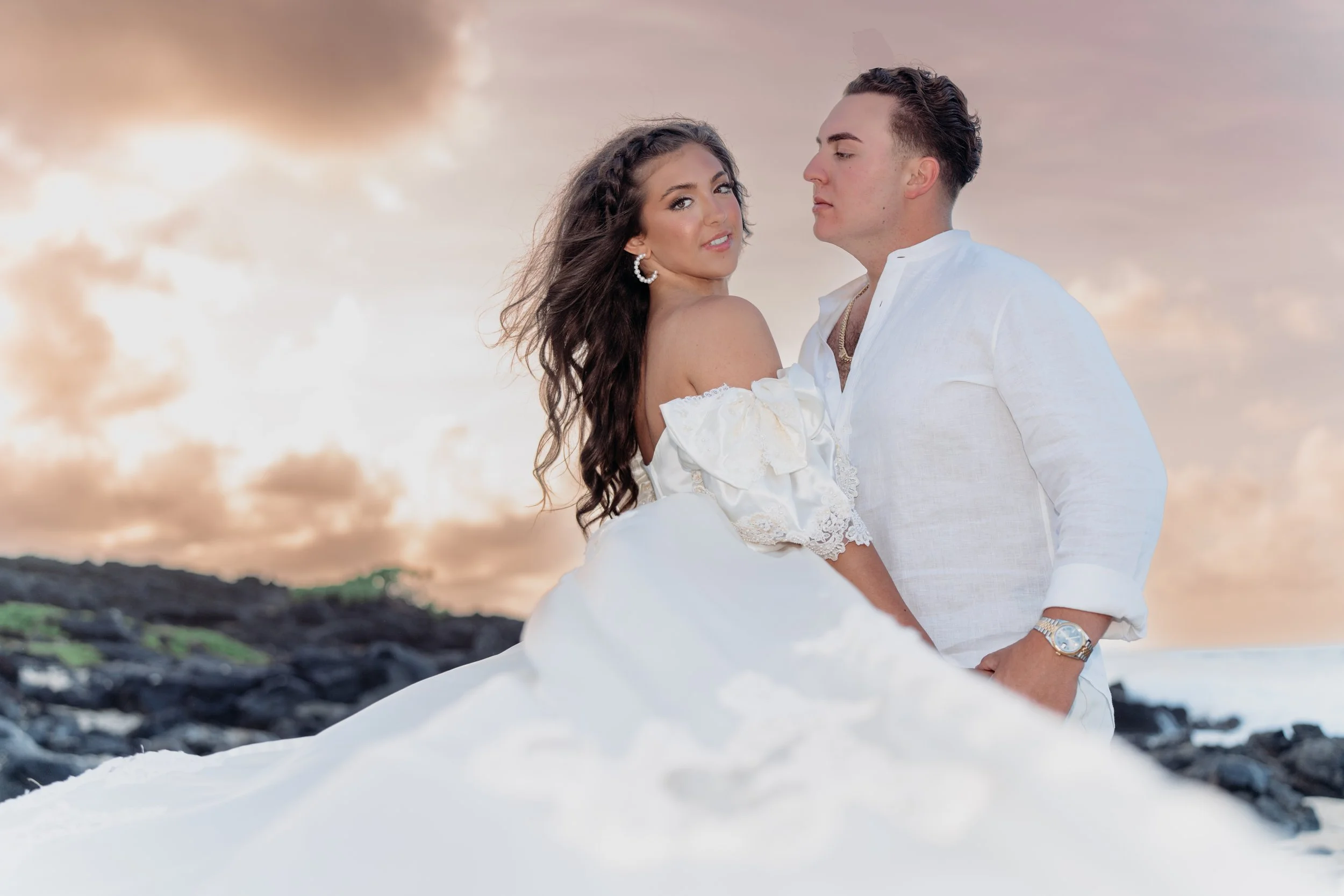 A couple in white clothing posing outdoors at sunset on a rocky beach, with the woman wearing an off-shoulder dress and the man in a white shirt, standing close together.