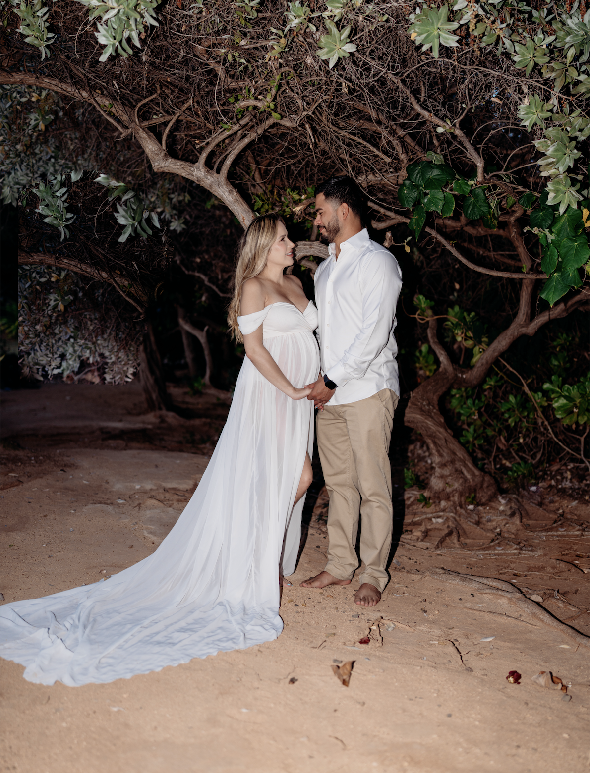 A couple holding hands in a romantic moment on a sandy beach at night, with a large tree with green leaves in the background.