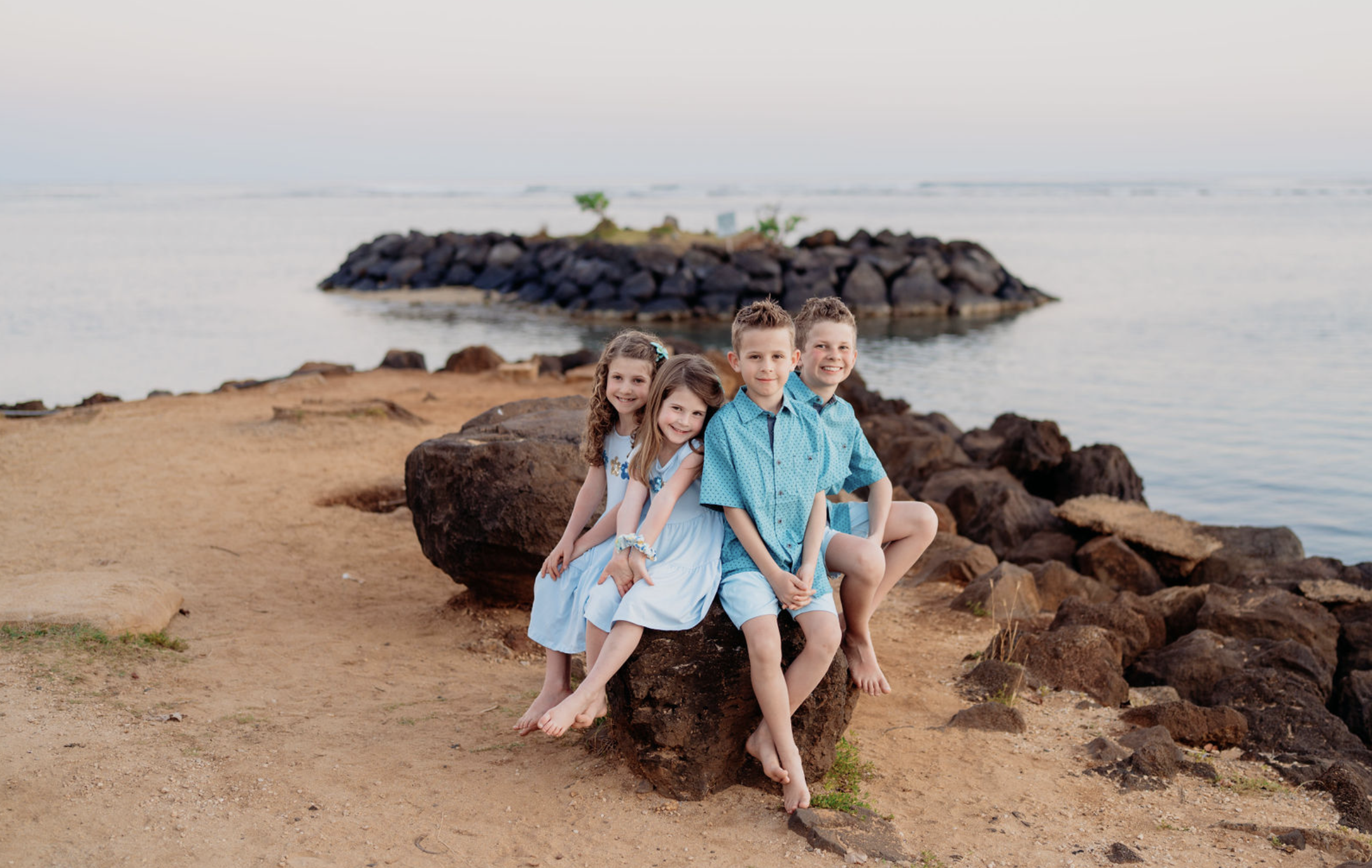 Four children in blue outfits sitting on a rock on a sandy beach with rocks and water in the background