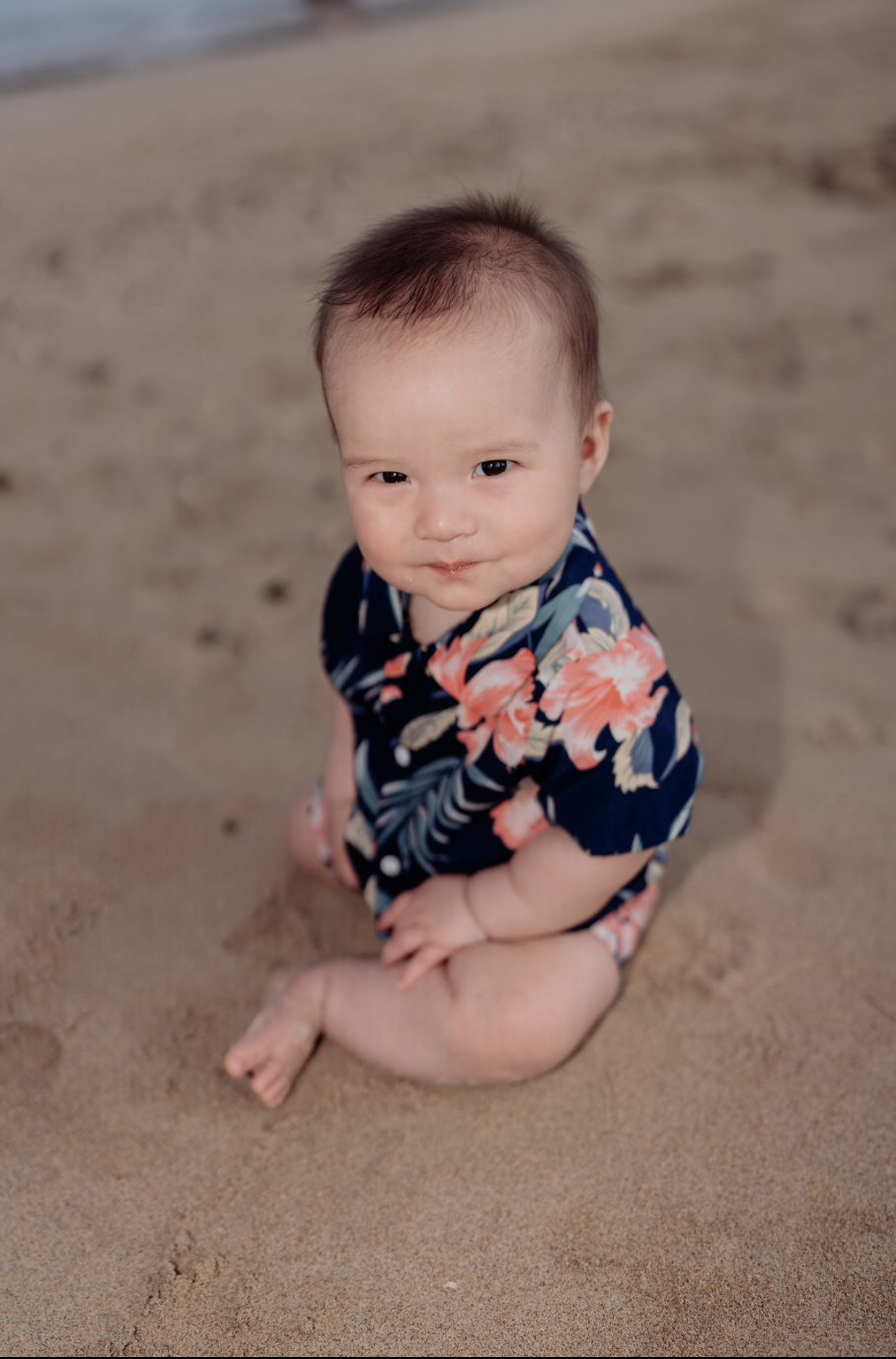 A young child with dark hair sitting on the sandy beach, wearing a floral navy blue shirt.