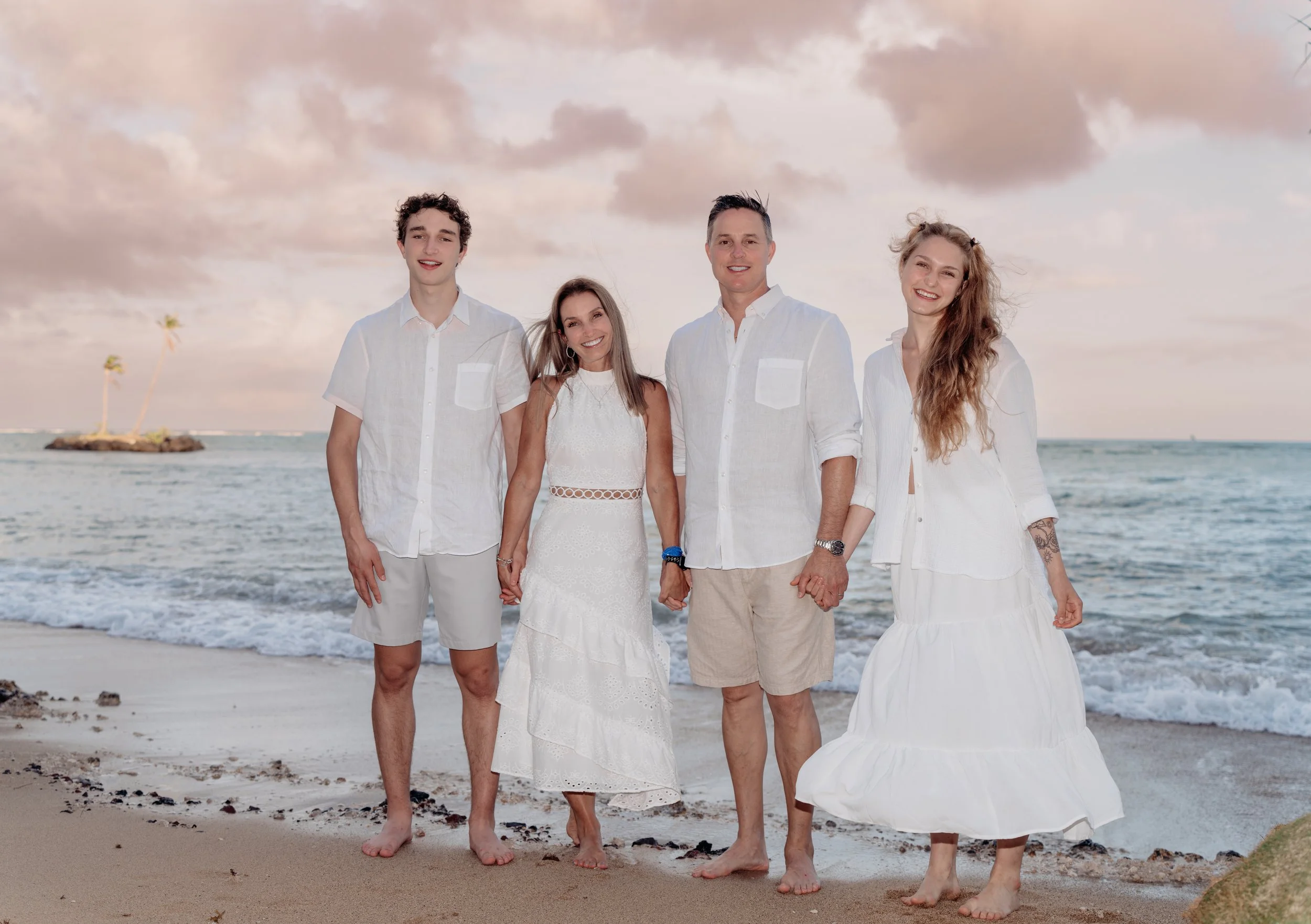 Family in white clothing walking on the beach with palm trees and ocean in the background.