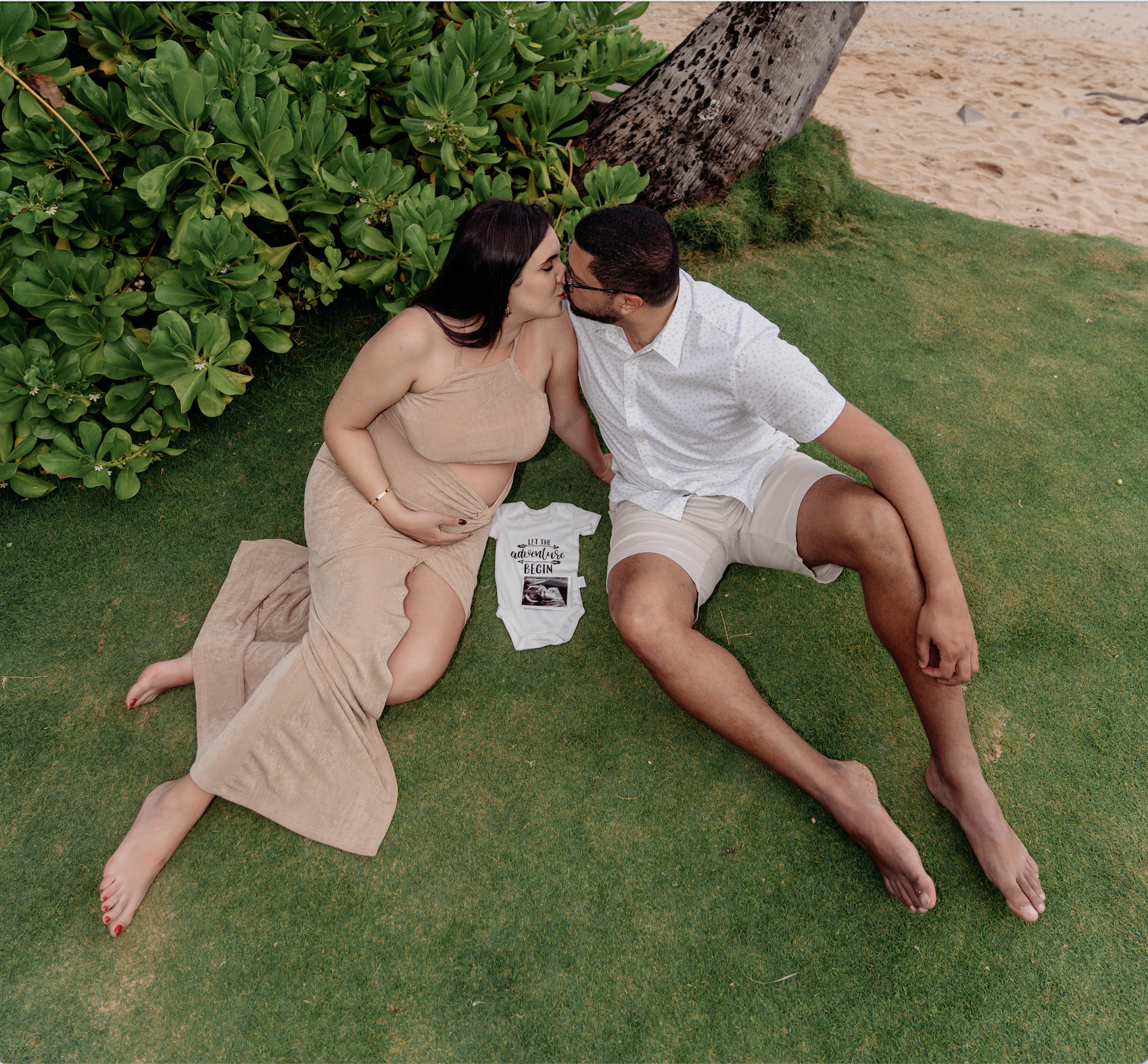 A couple sitting on grass, kissing, with a pregnant woman. The woman is in a beige dress, and the man is in a white shirt and beige shorts. A baby onesie with the text 'Let the adventure begin' and a photo is placed between them. They are near a tree