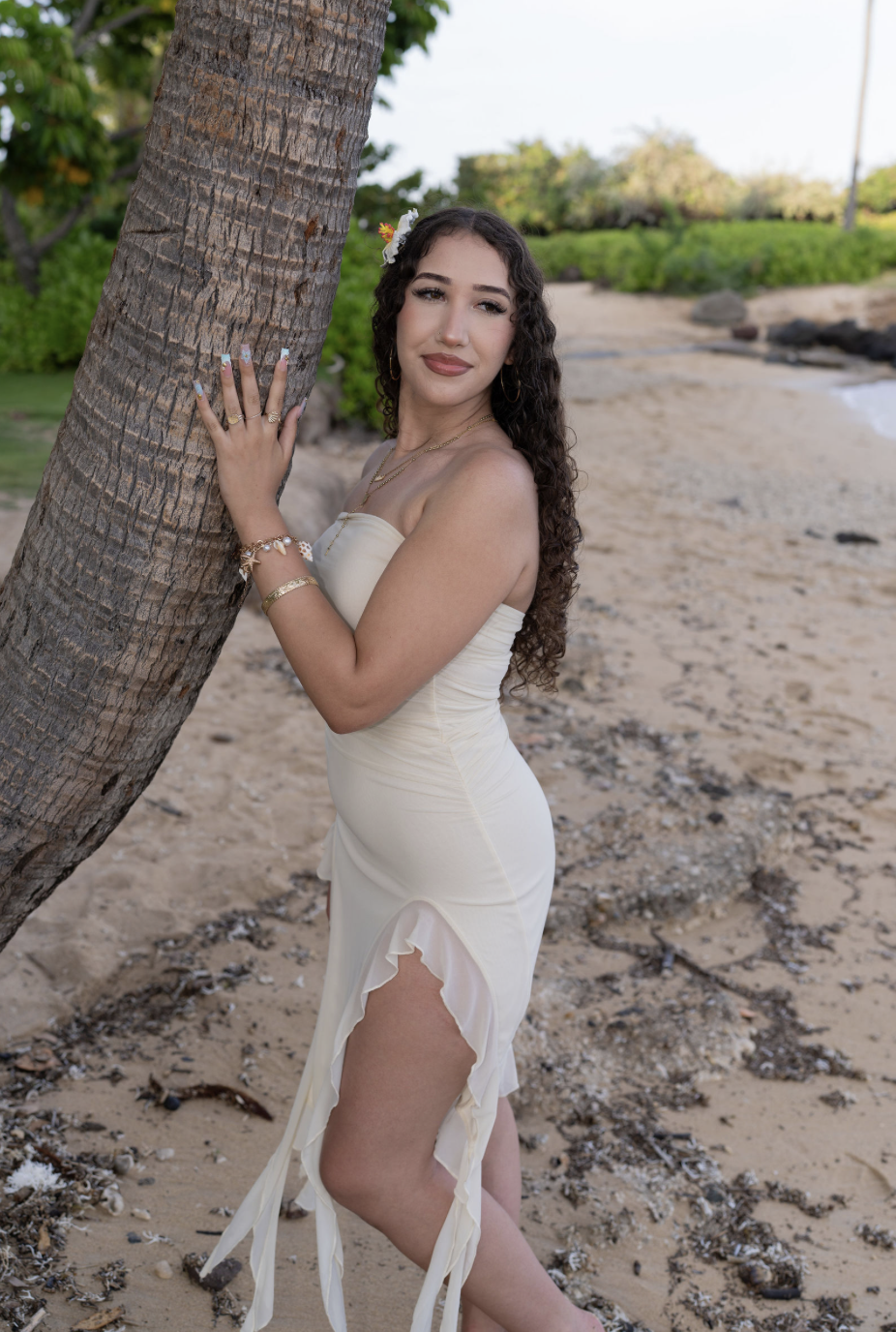 A woman in a white dress with ruffles on the side, standing on a sandy beach with one hand on a palm tree, looking at the camera with a slight smile.