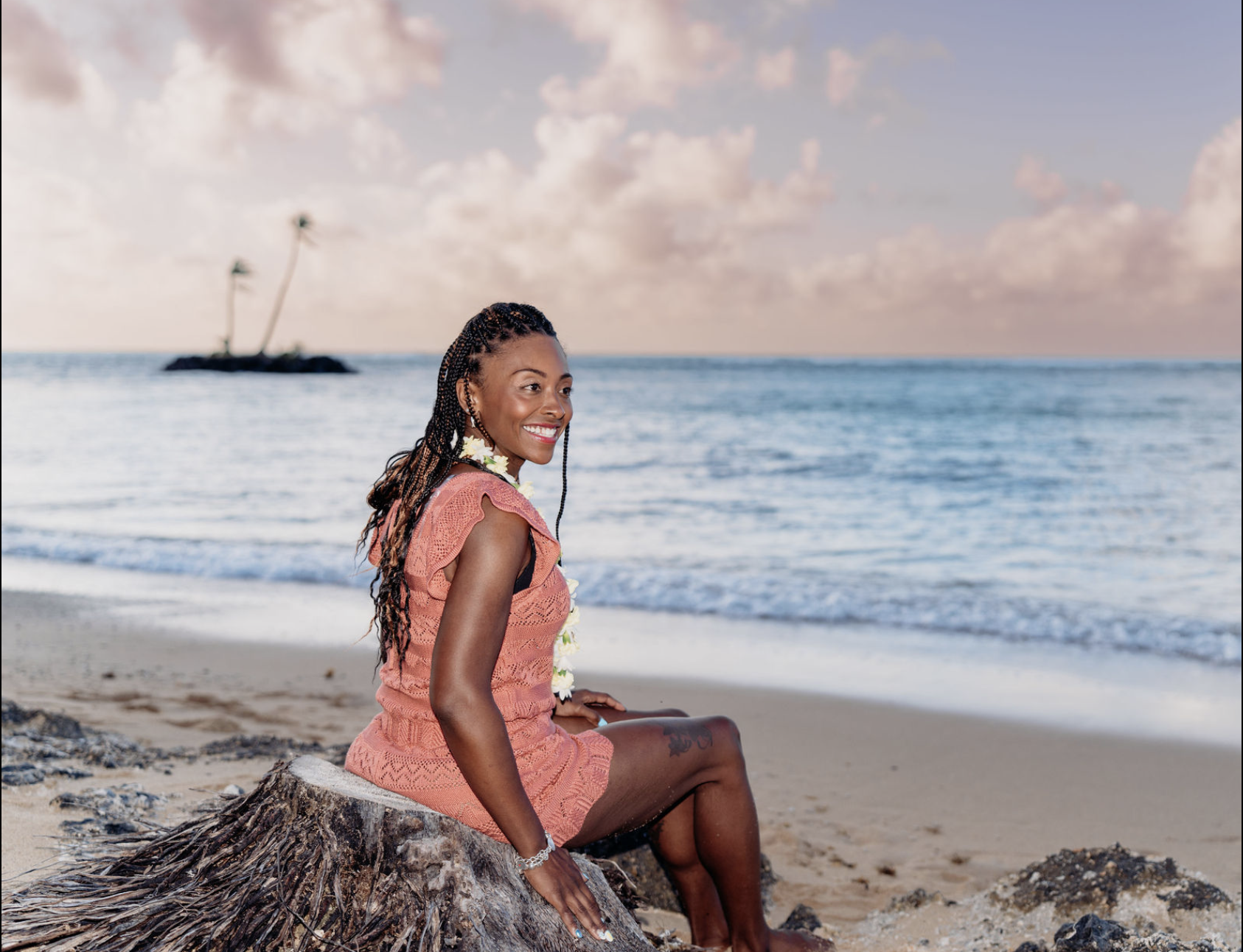 A smiling woman with braided hair in a pink dress sitting on a tree trunk on a beach with the ocean and a small island with palm trees in the background during sunset.