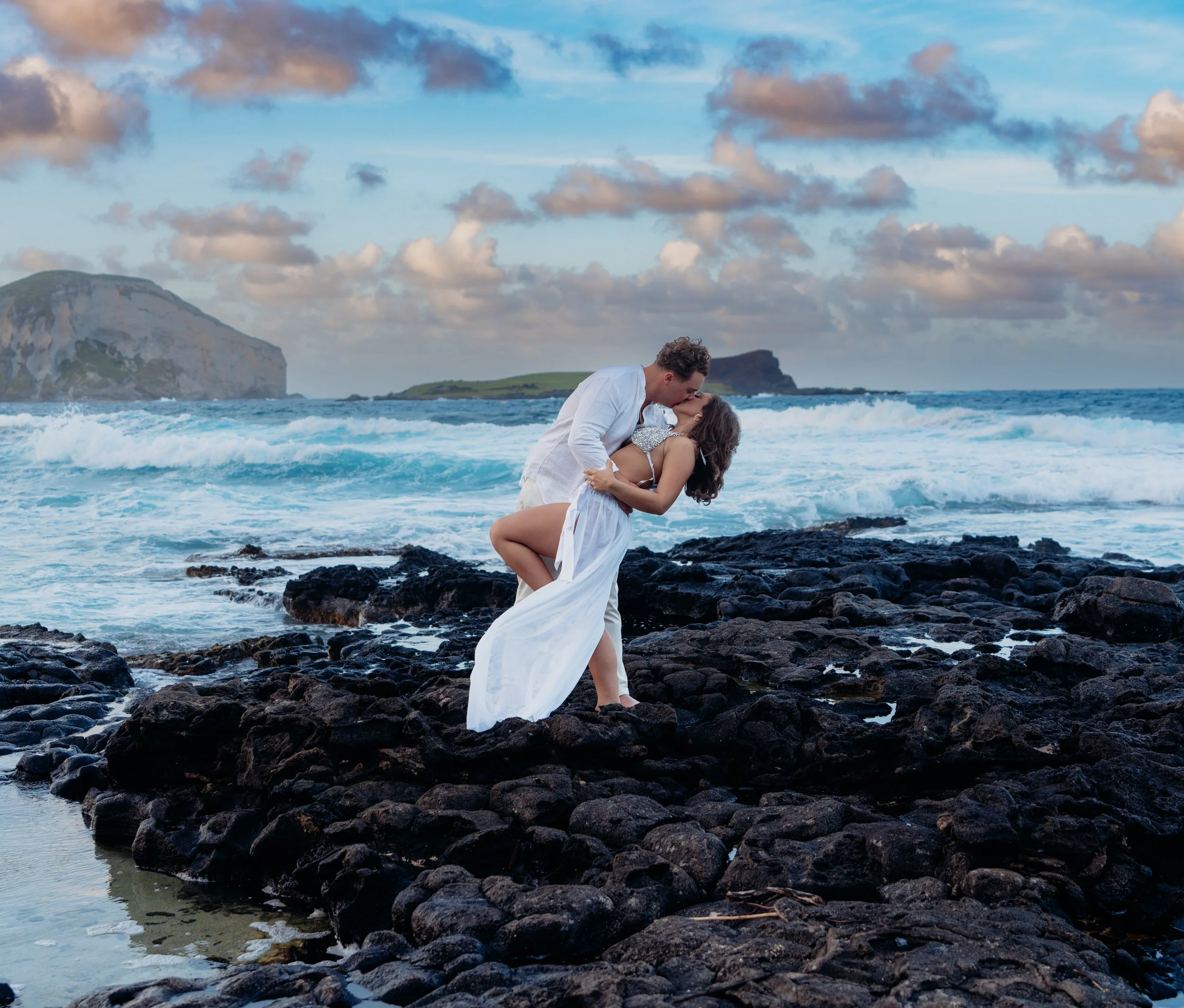 A couple dressed in white clothes sharing a romantic kiss on black rocks by the ocean with waves crashing, ocean cliffs in the background, under a partly cloudy sky.