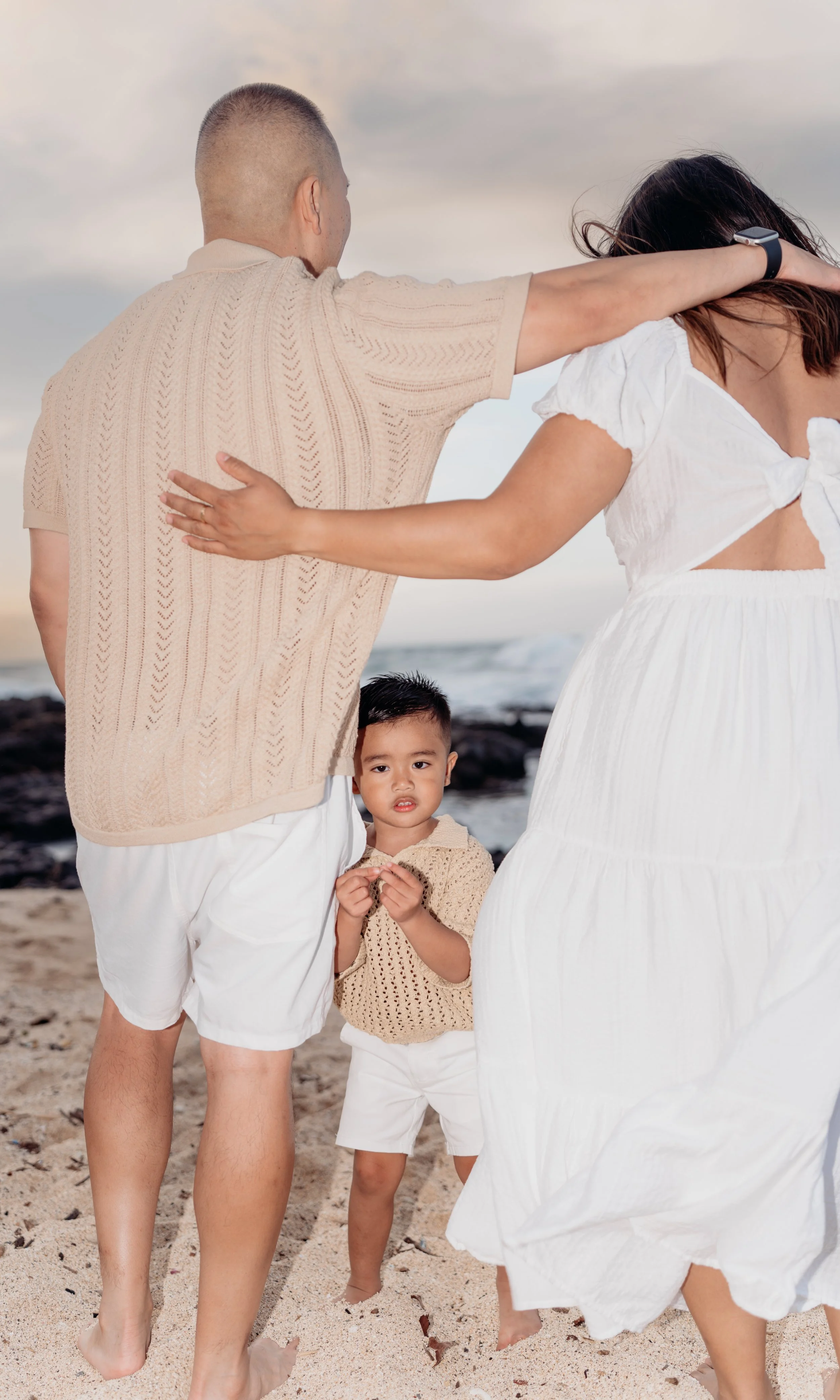 Family on the beach, a man with a shaved head in a beige sweater, woman in a white dress, and a young boy in a beige sweater, with a rocky shoreline and ocean in the background.