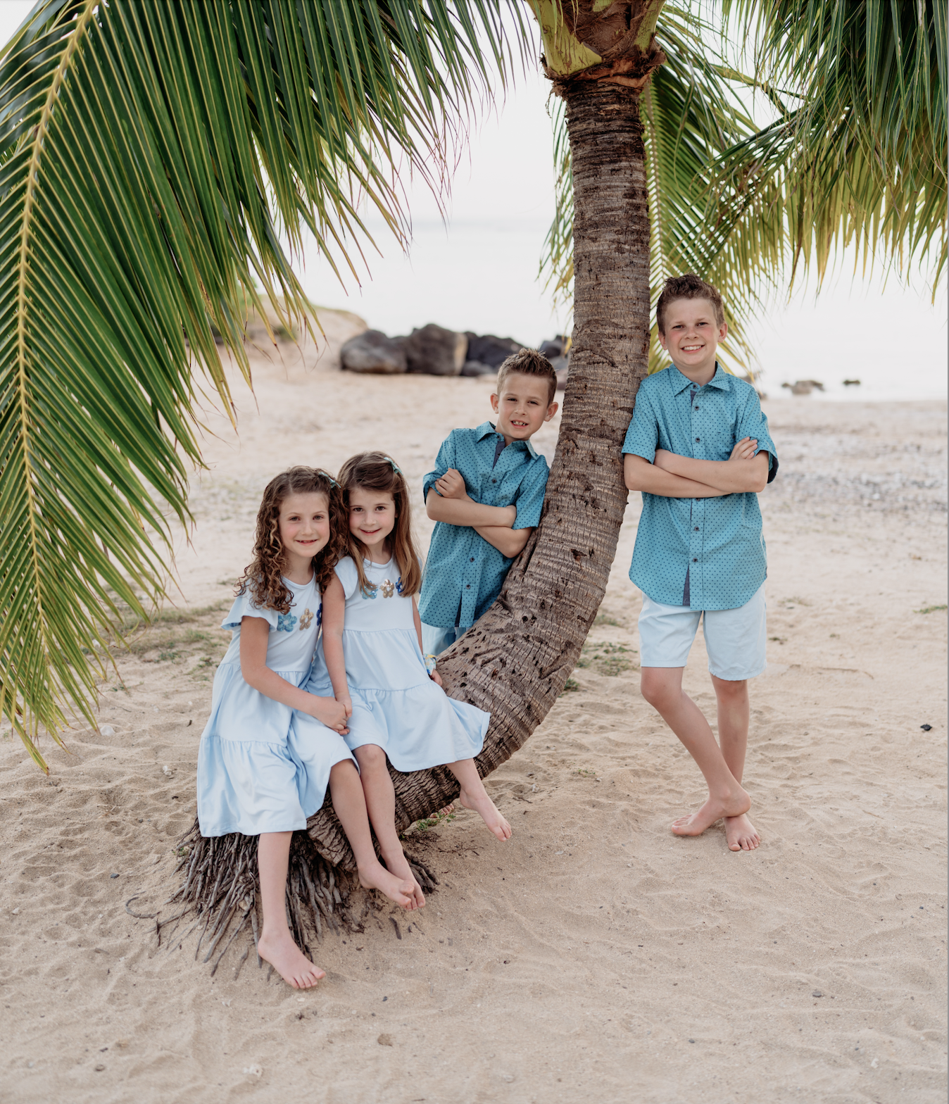 Four children, two girls and two boys, are posing on a sandy beach under a palm tree, wearing light blue and teal clothing, smiling at the camera.