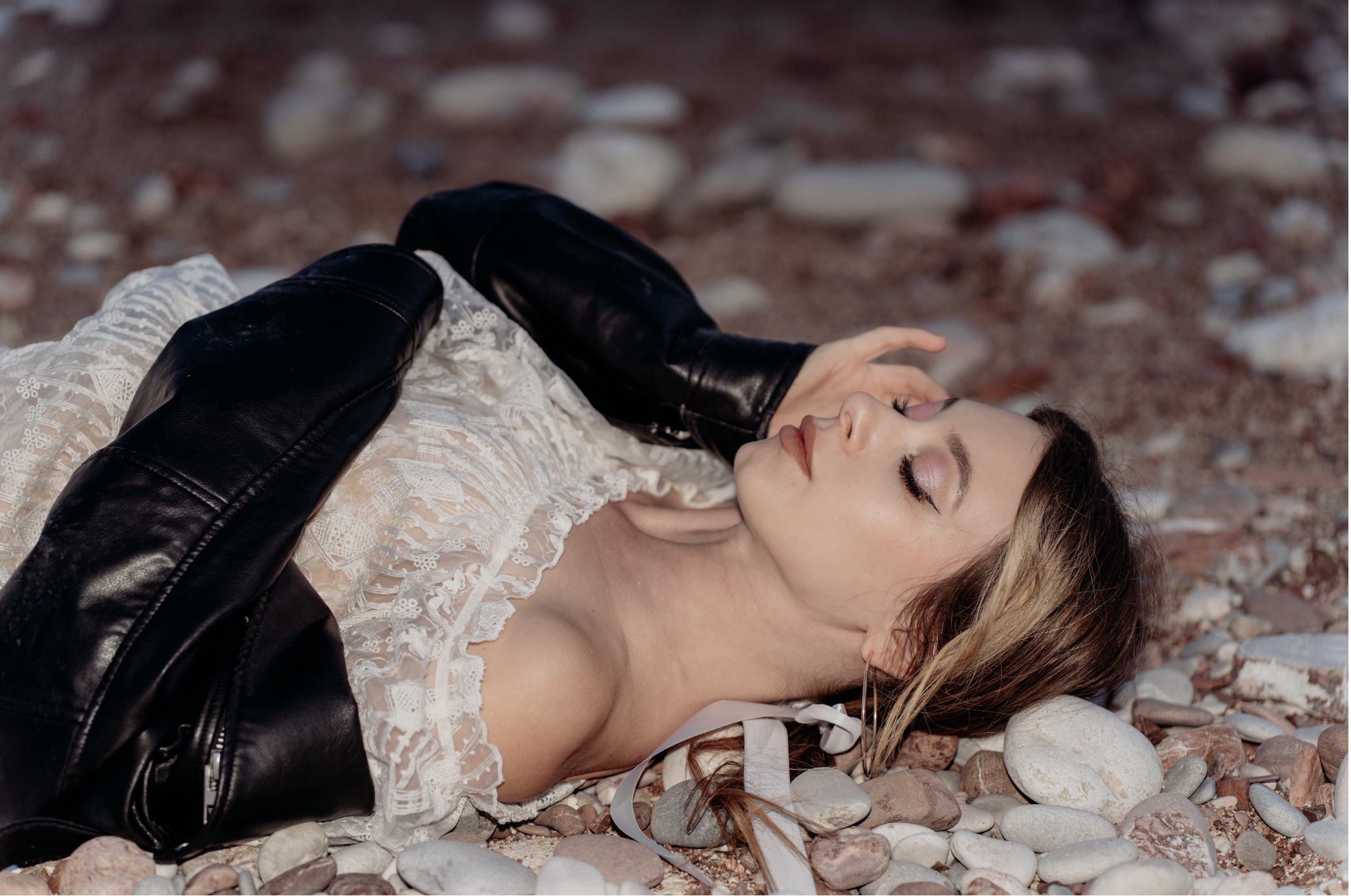 A woman lying on a rocky beach, wearing a lace dress and a leather jacket, with her eyes closed and hair spread out on the stones.