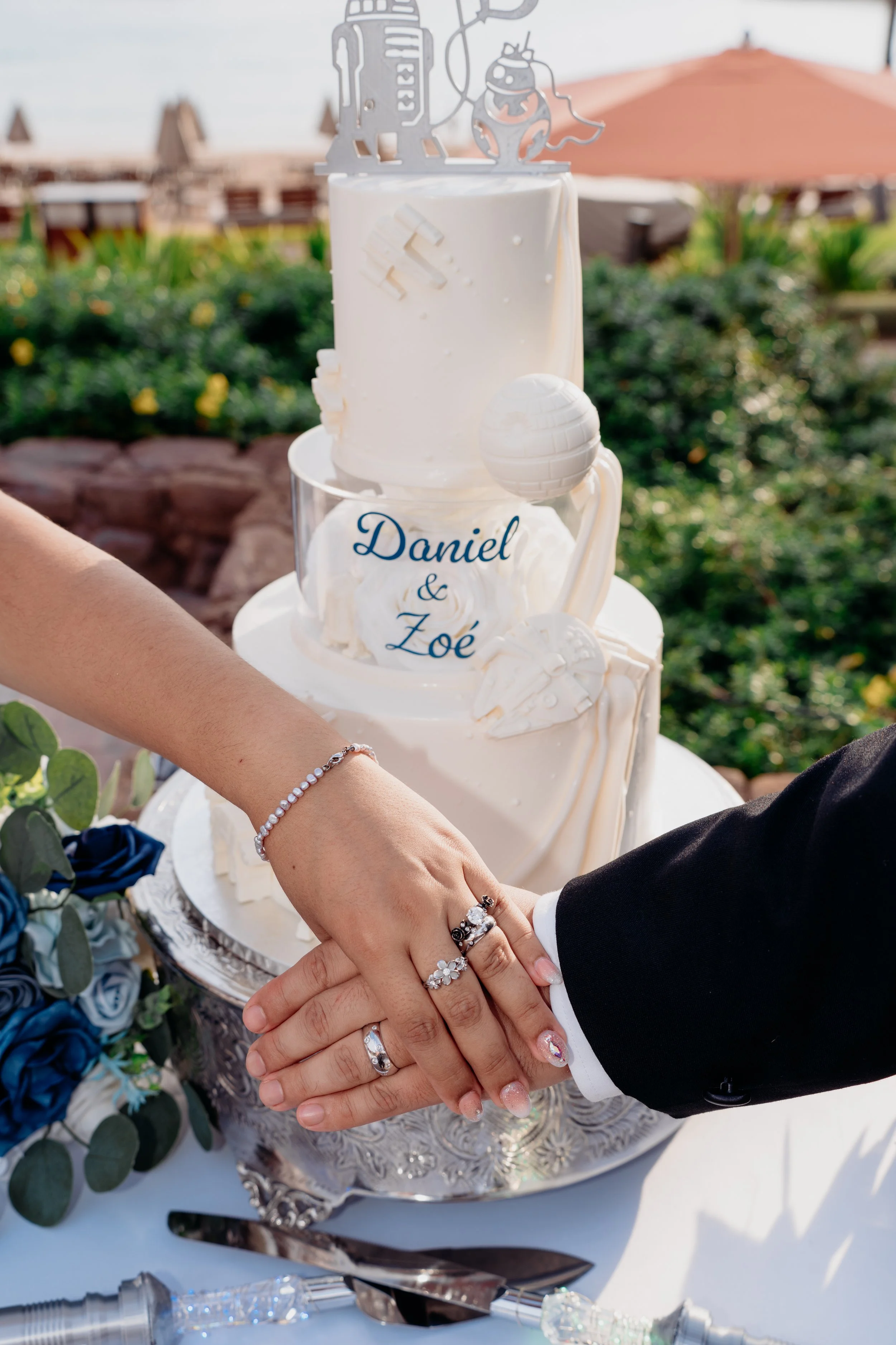 A wedding cake with white frosting, topped with space-themed decorations, and the names 'Daniel & Zoé' written on a piece of fondant, with a bride and groom's hands placed on top of the cake in a celebratory gesture.