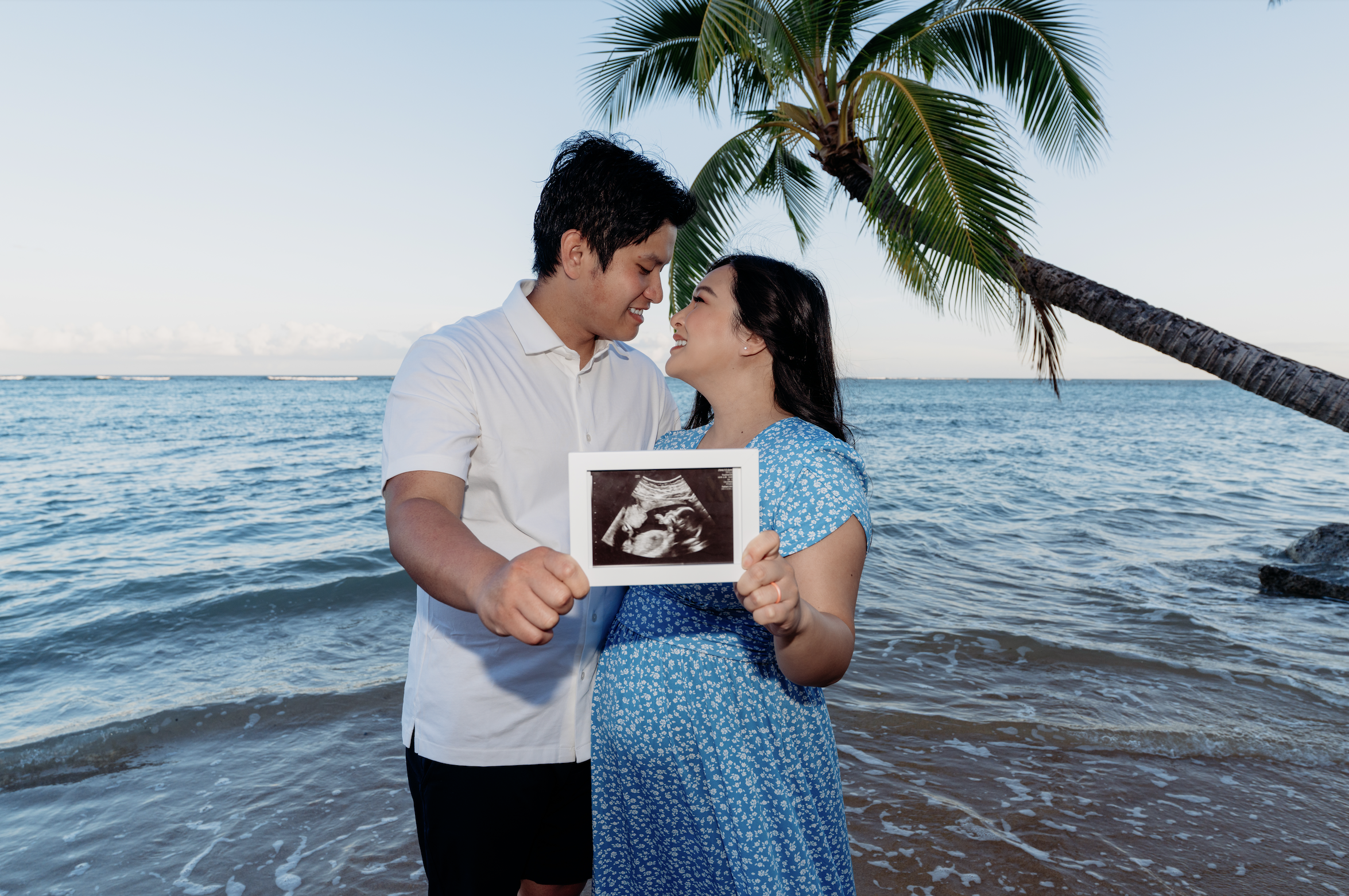 A couple holding an ultrasound picture at the beach with a palm tree and ocean in the background.
