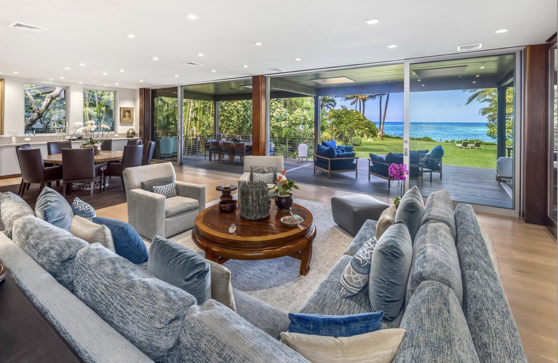 Spacious living room with large sliding glass doors opening to a porch and a tropical ocean view, furnished with a large L-shaped sofa, armchairs, a round wooden coffee table, and a dining area.