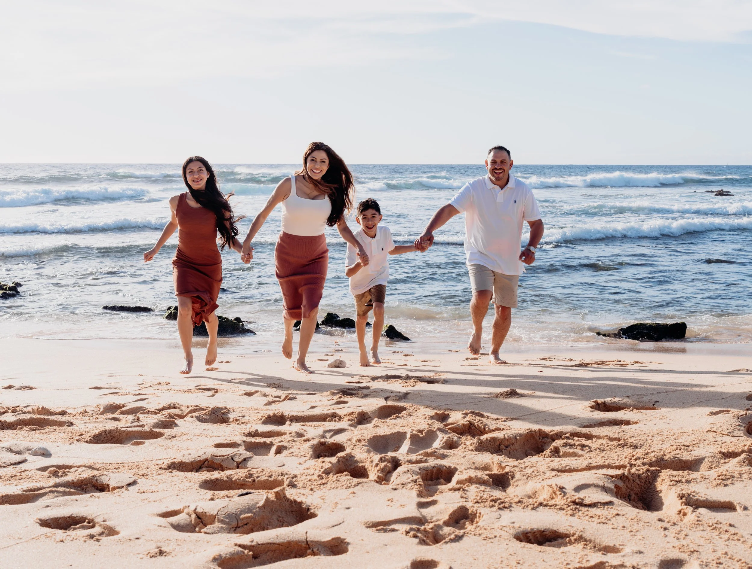 Family of four walking hand-in-hand on the beach near the ocean, with footprints in the sand and waves in the background.