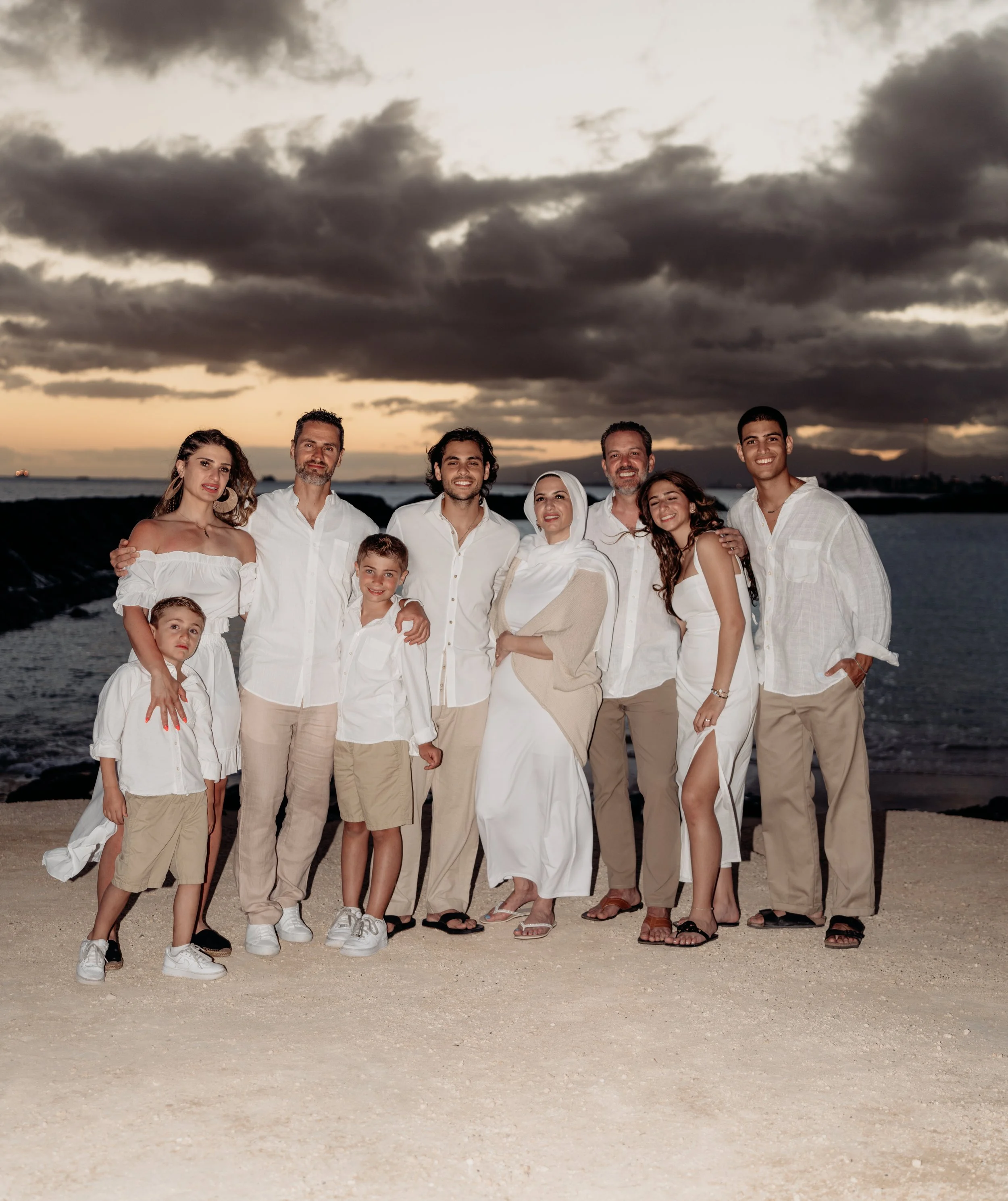 A multi-generational family dressed in white and beige standing on a beach at sunset with dark clouds overhead.