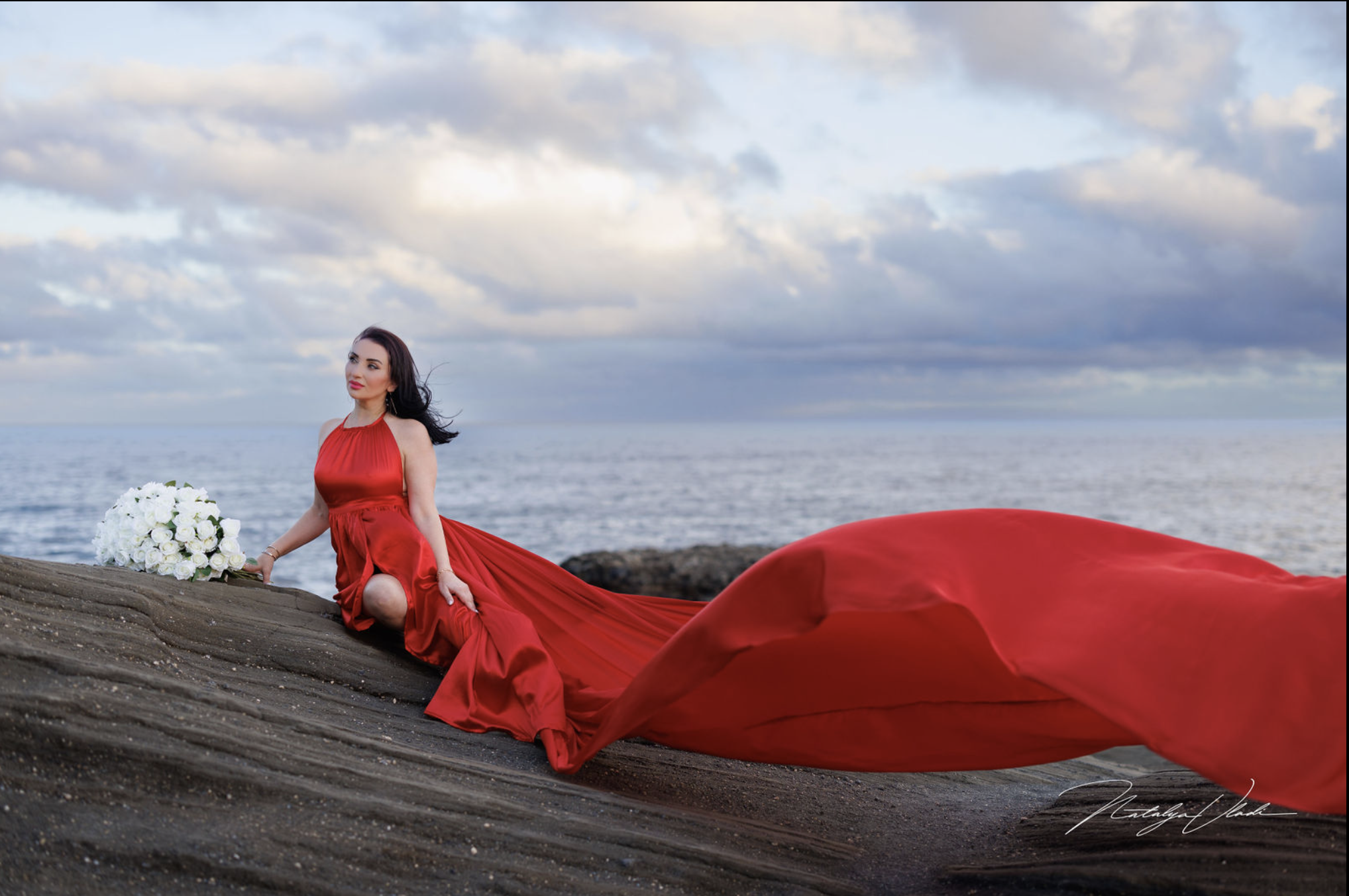 A woman in a flowing red dress kneeling on a rocky shoreline near the ocean with a bouquet of white roses and a cloudy sky in the background.