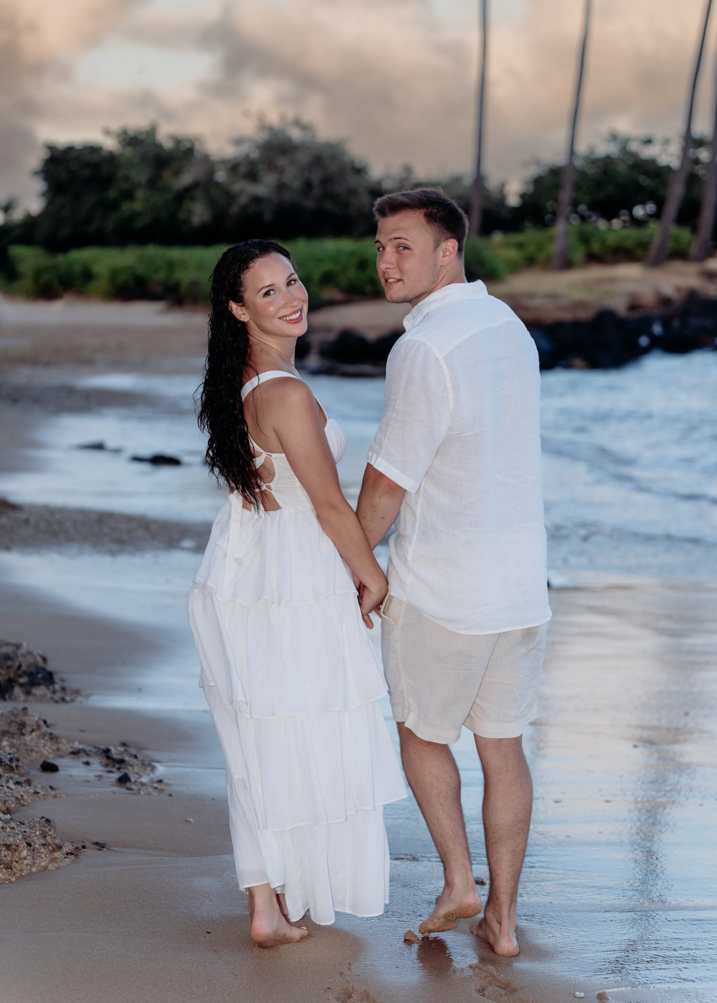 A couple holding hands on the beach during sunset, with palm trees and ocean waves in the background.