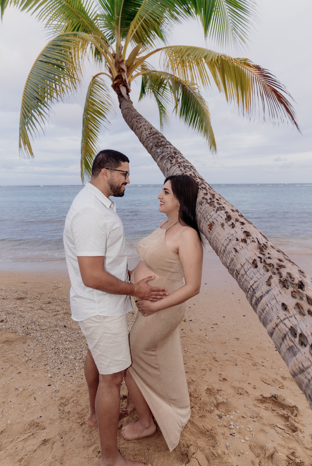 A couple standing on a sandy beach by the ocean, with a leaning palm tree above them. The man and woman are gazing at each other, smiling, and the woman is holding her pregnant belly. The scene is tropical and serene.