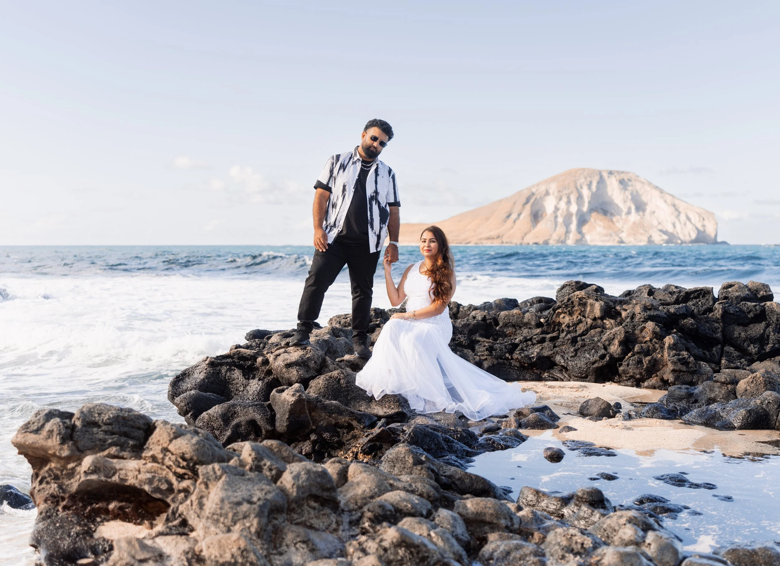 A couple on a rocky beach; the woman in a white dress is seated on rocks and holding the man's hand, who is standing, dressed casually, with an ocean and a distant island in the background.