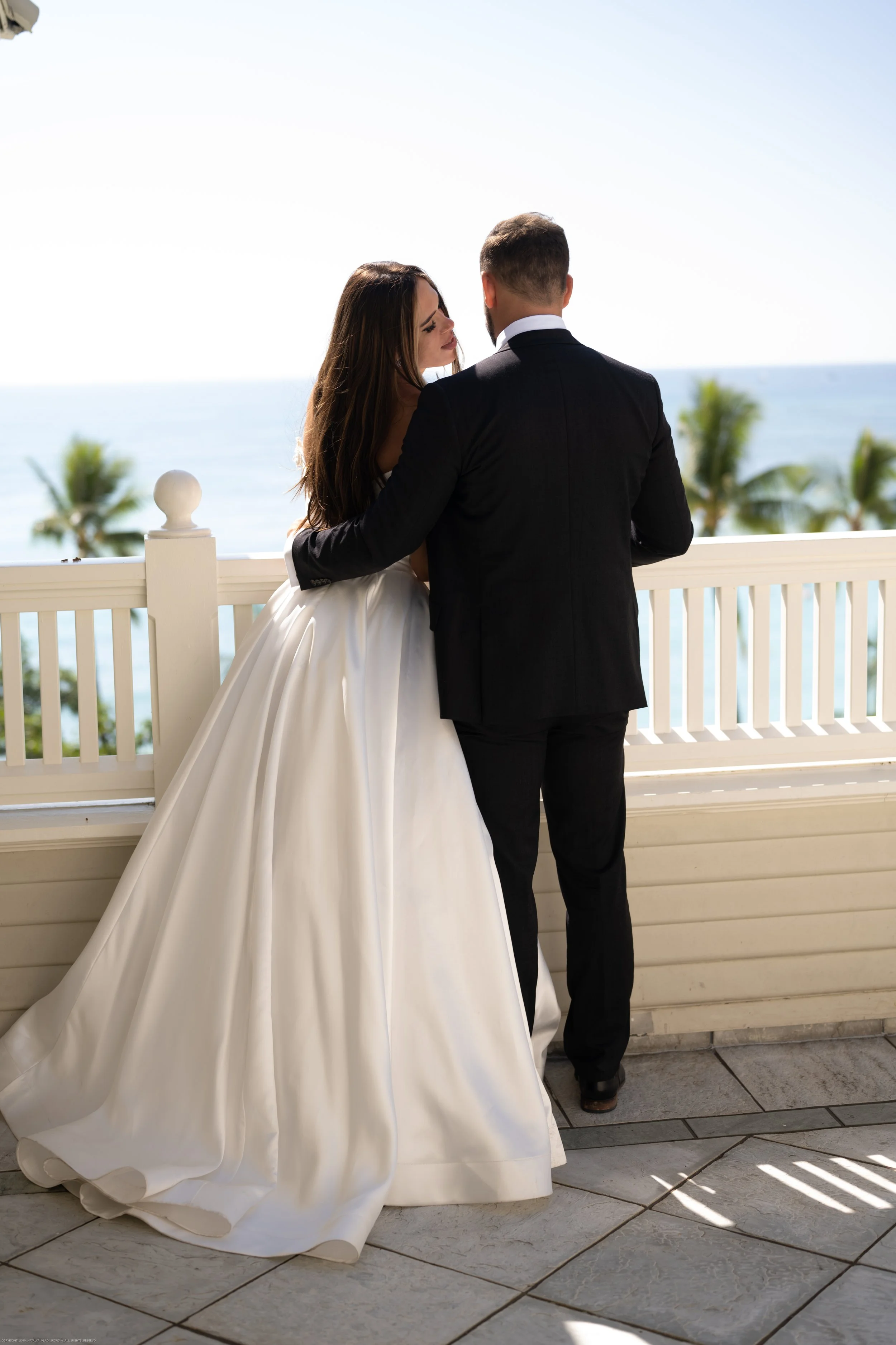 A bride and groom on a balcony with ocean view, the bride is in a white wedding gown, and the groom in a black suit.
