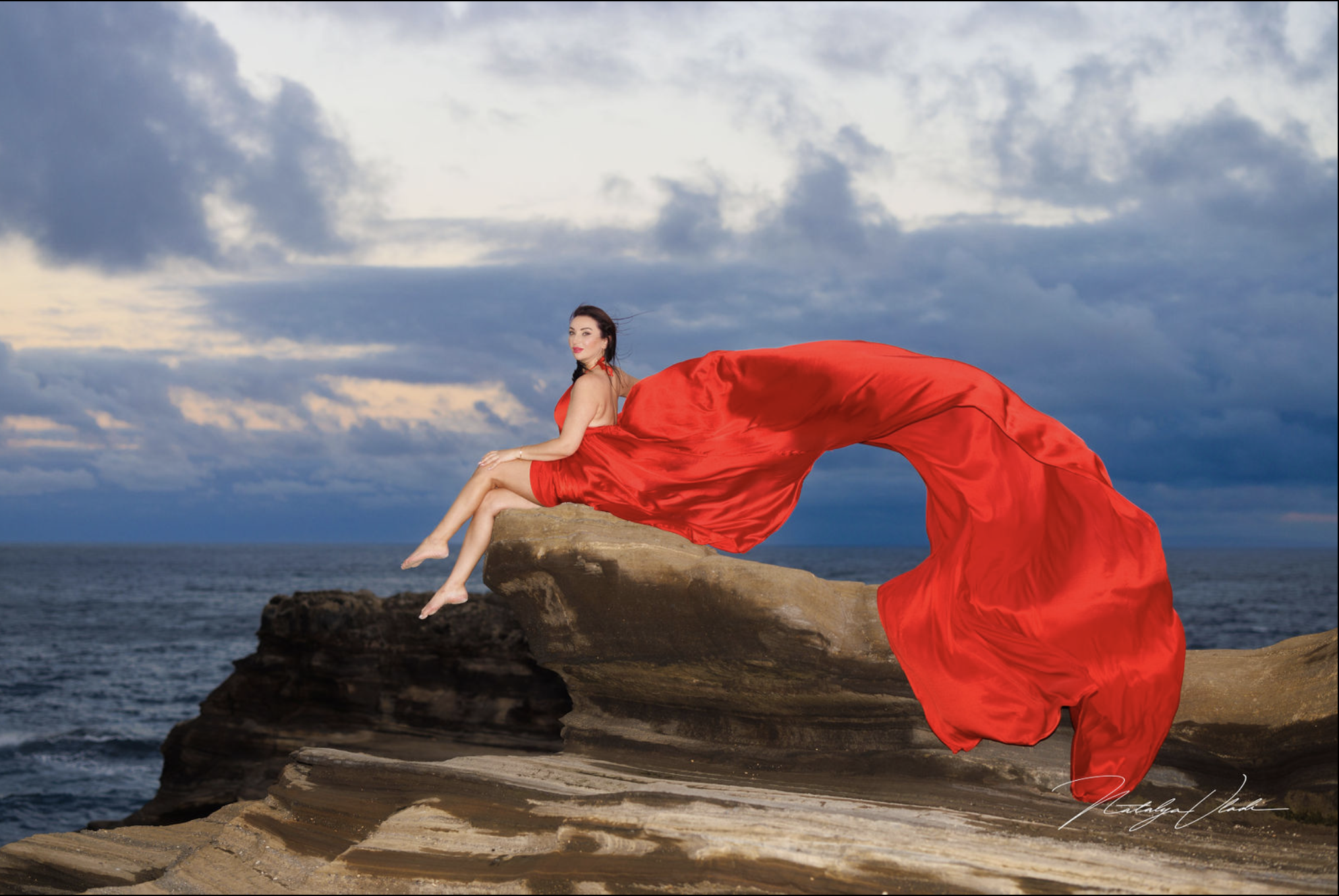 A woman in a flowing red dress is sitting on a rock formation by the ocean with a cloudy sky in the background.