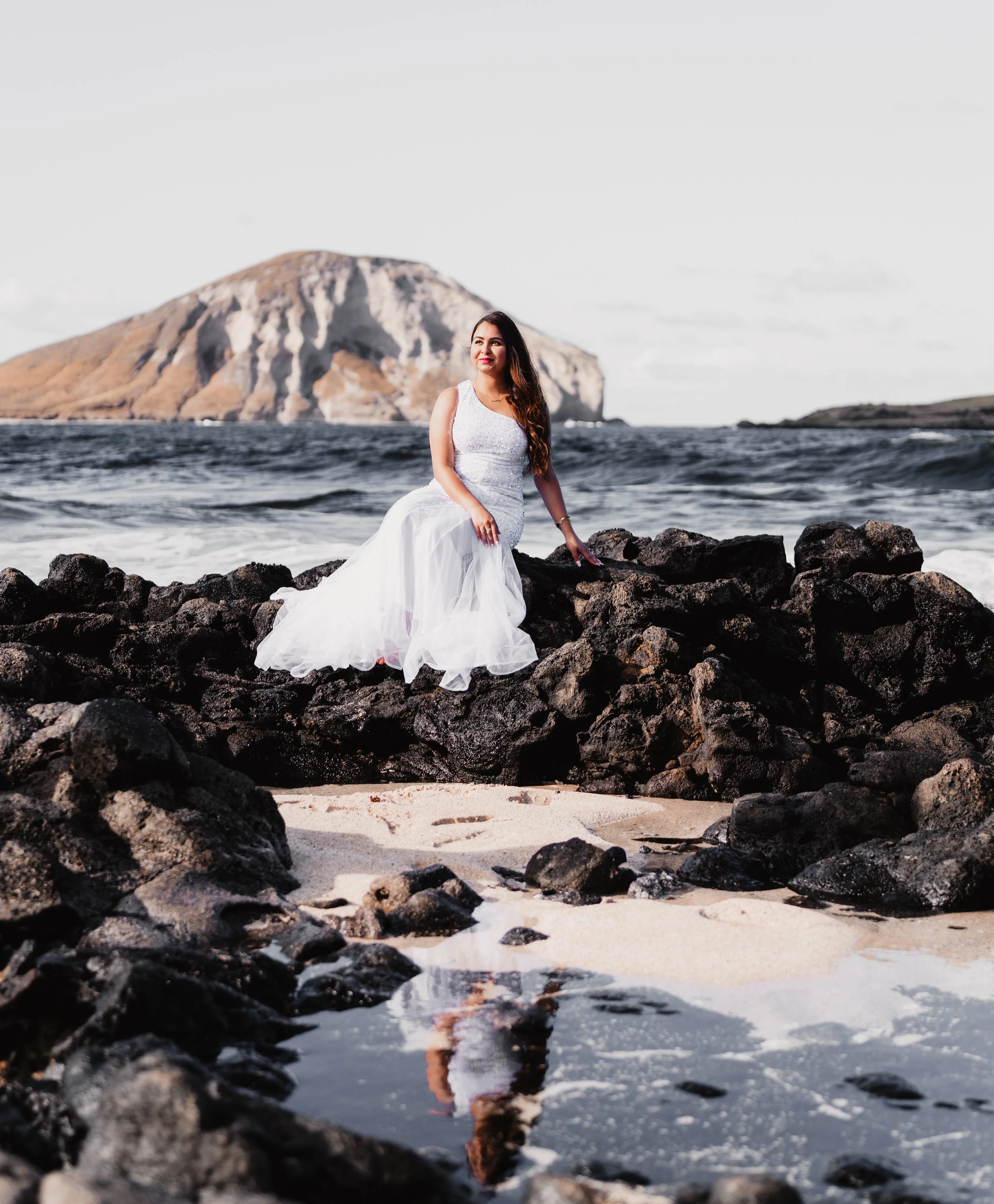 Woman in wedding dress sitting on rocks on a beach with ocean waves and a mountain in the background