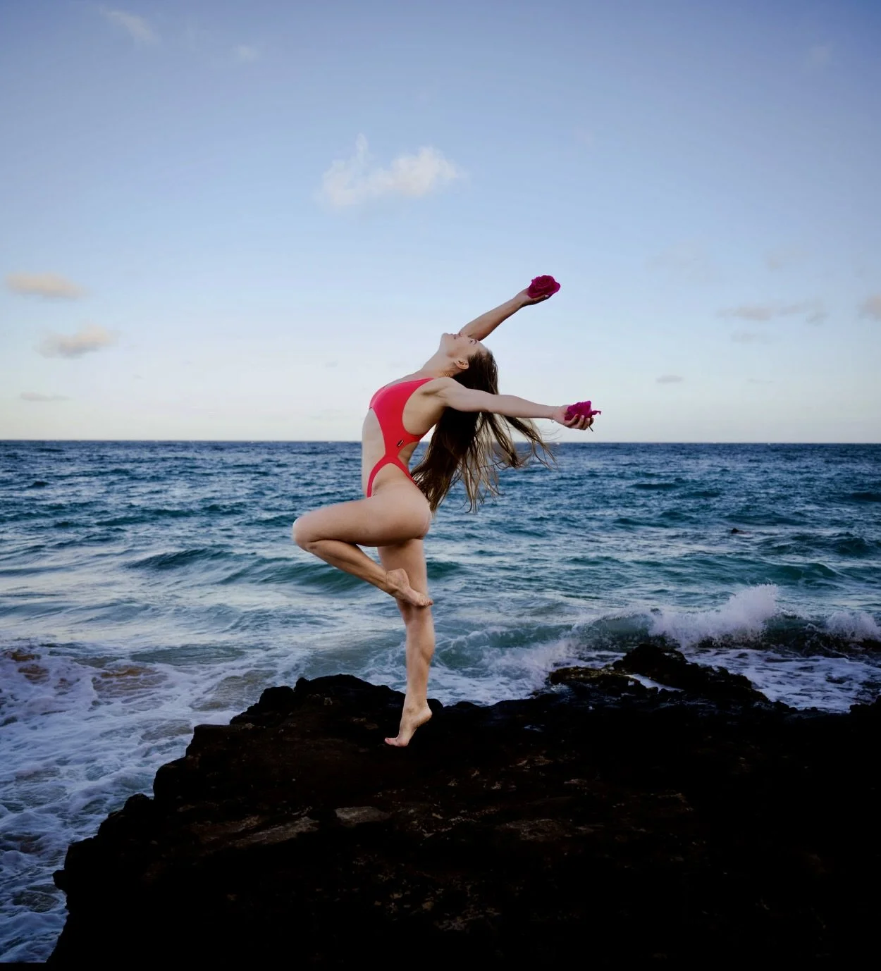 A woman in a red swimsuit performing a yoga pose on rocks by the ocean, holding pink flowers, with a blue sky and scattered clouds in the background.