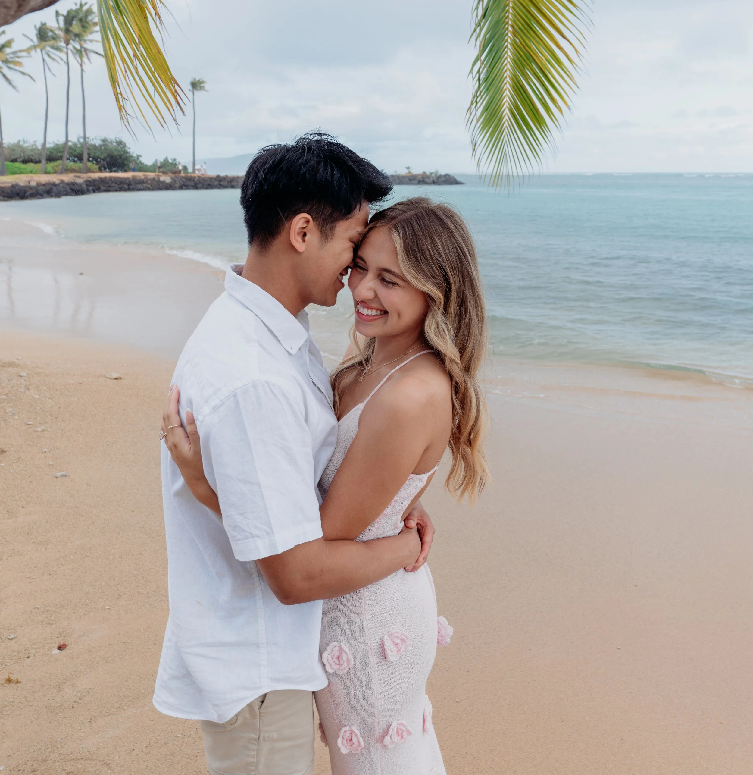 A happy couple embracing and smiling on a beach with palm trees in the background.