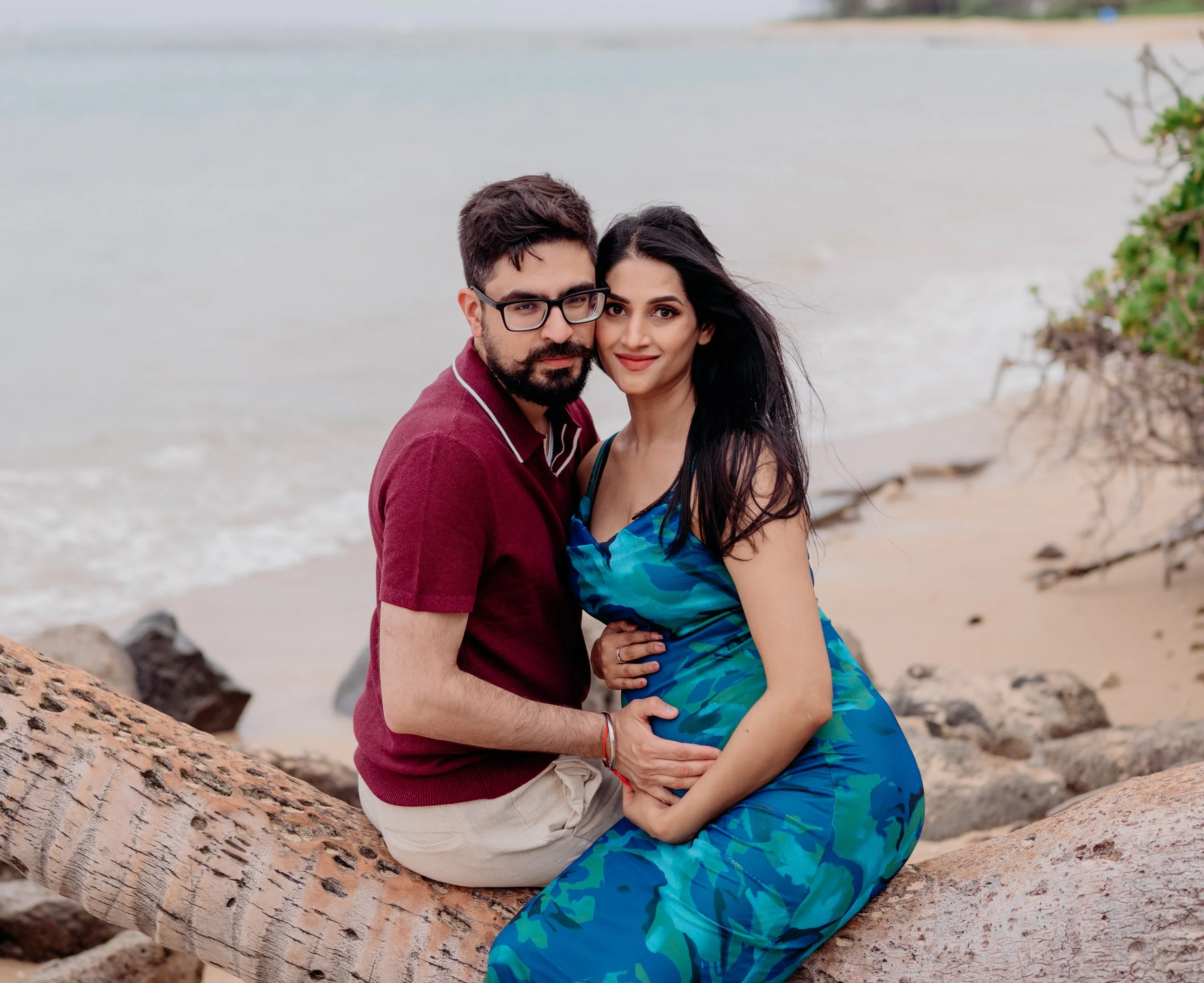 A couple sitting on a large driftwood log on the beach, embracing each other with the ocean and rocks in the background, during daytime.
