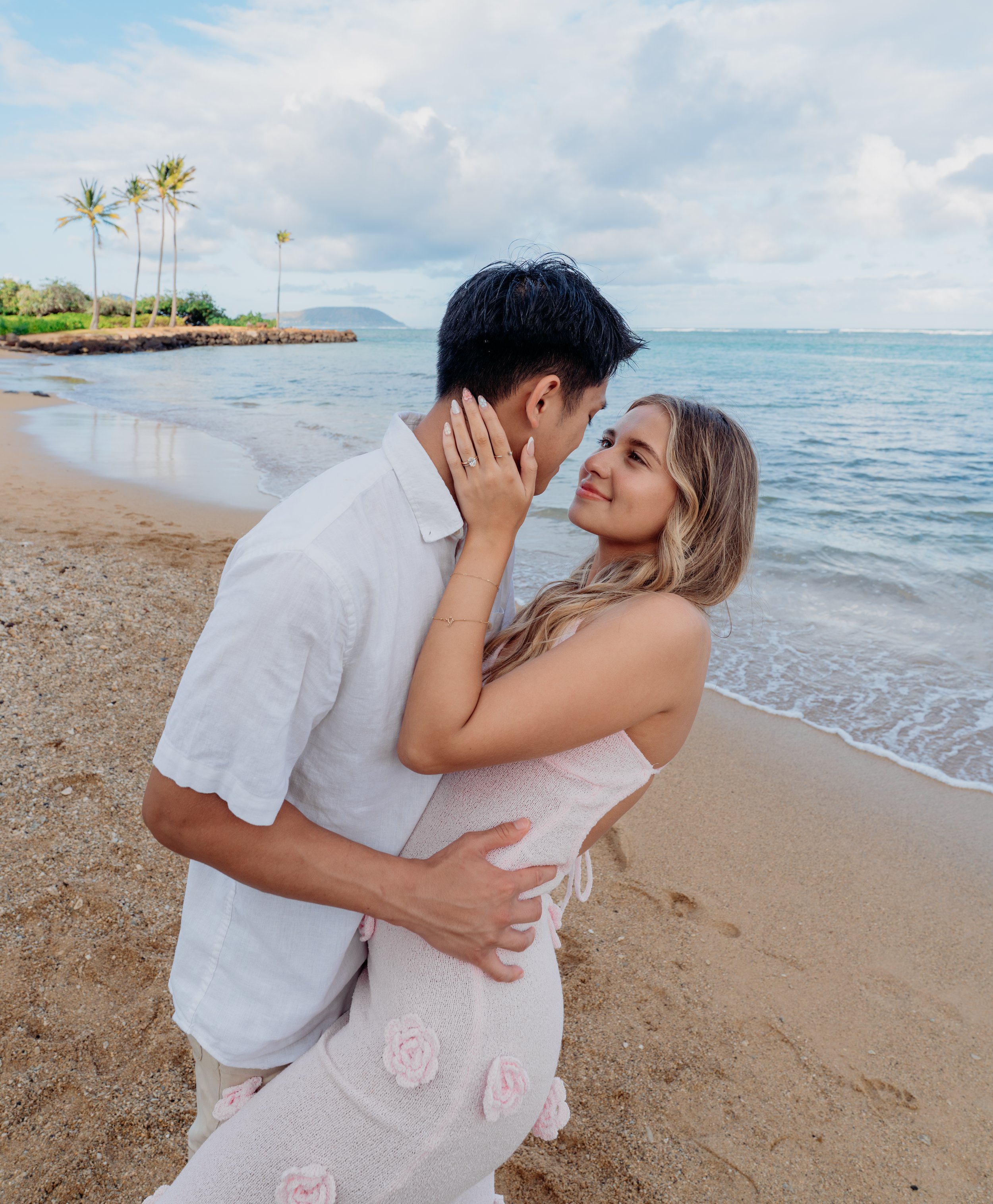 A couple embracing on a beach with palm trees, sandy shore, and ocean in the background.