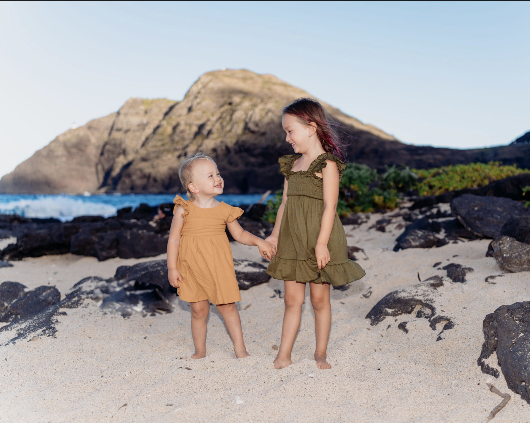 Two young girls in matching dresses holding hands and smiling on a sandy beach with rocks, ocean, and a mountainous landscape in the background.