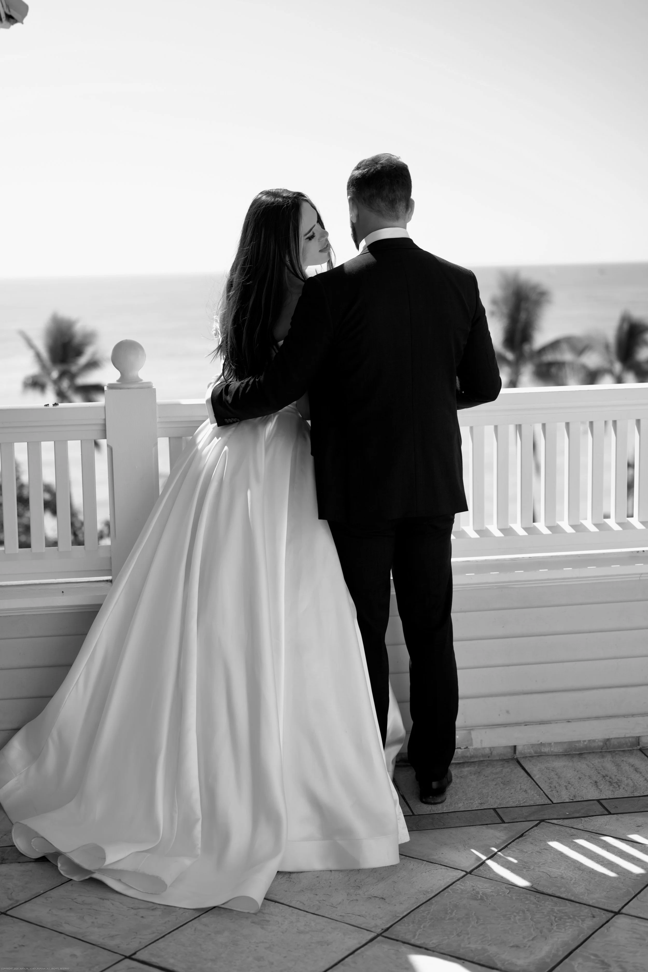 A bride and groom standing on a balcony, facing each other, with the ocean and palm trees in the background, black and white photograph.