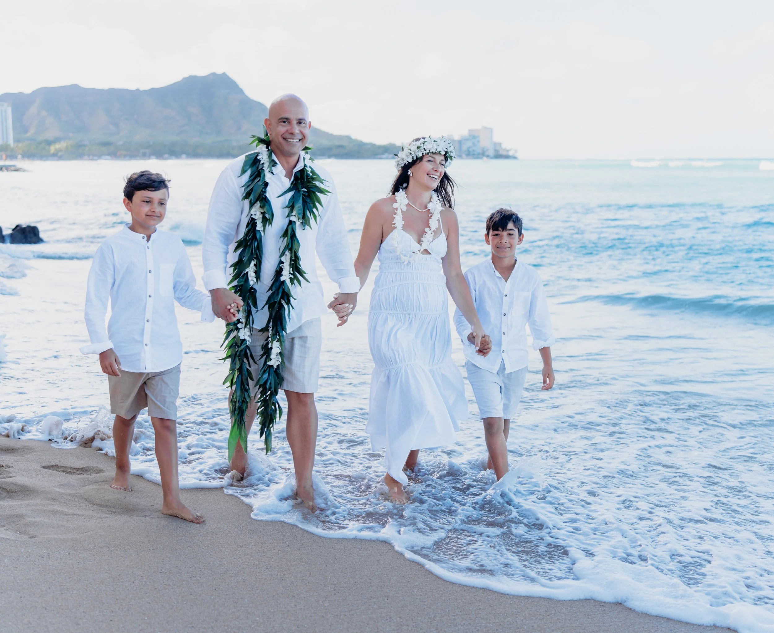 A couple getting married on a beach with two children, holding hands, all dressed in white, with ocean and mountains in the background.