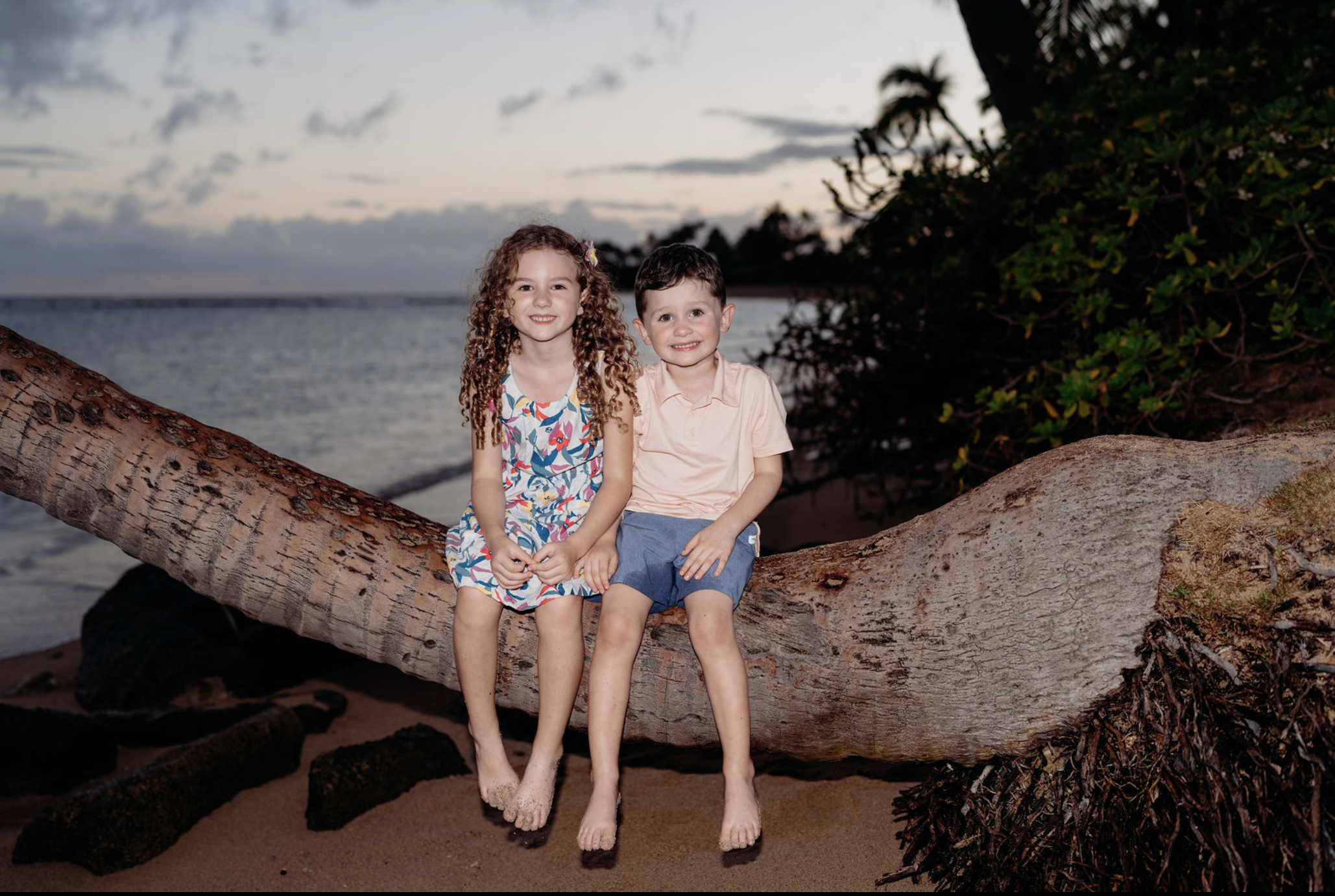 Two young children, a girl with curly hair wearing a colorful dress and a boy wearing a pink shirt and blue shorts, sitting on a large fallen tree trunk on the beach at sunset.