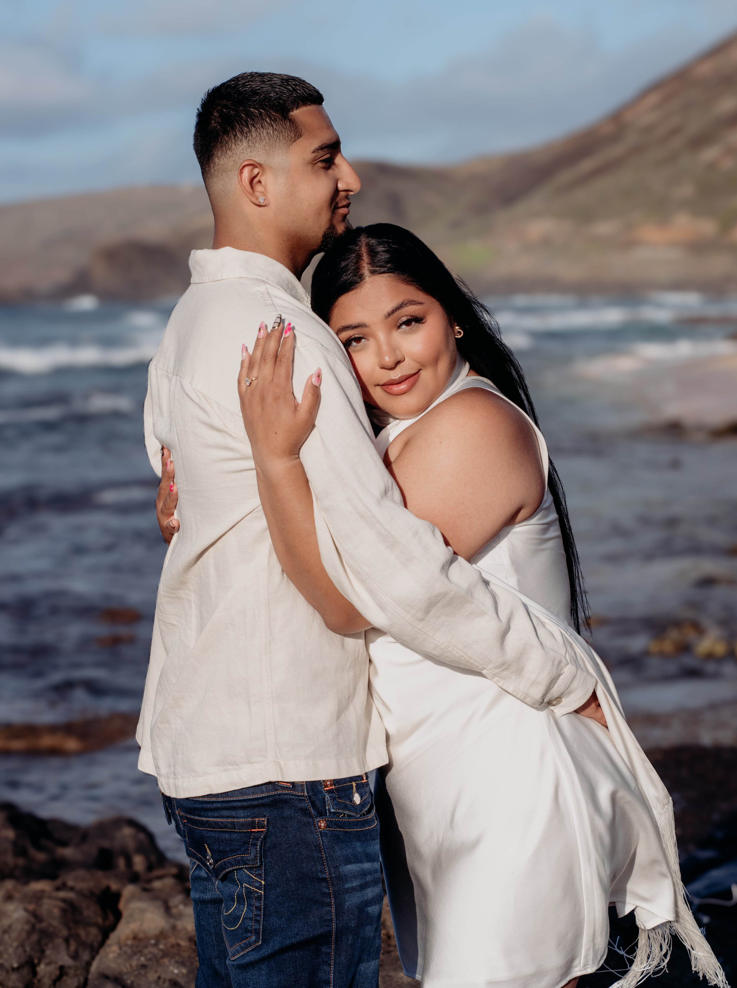 A couple embracing near the beach with ocean waves and hills in the background.