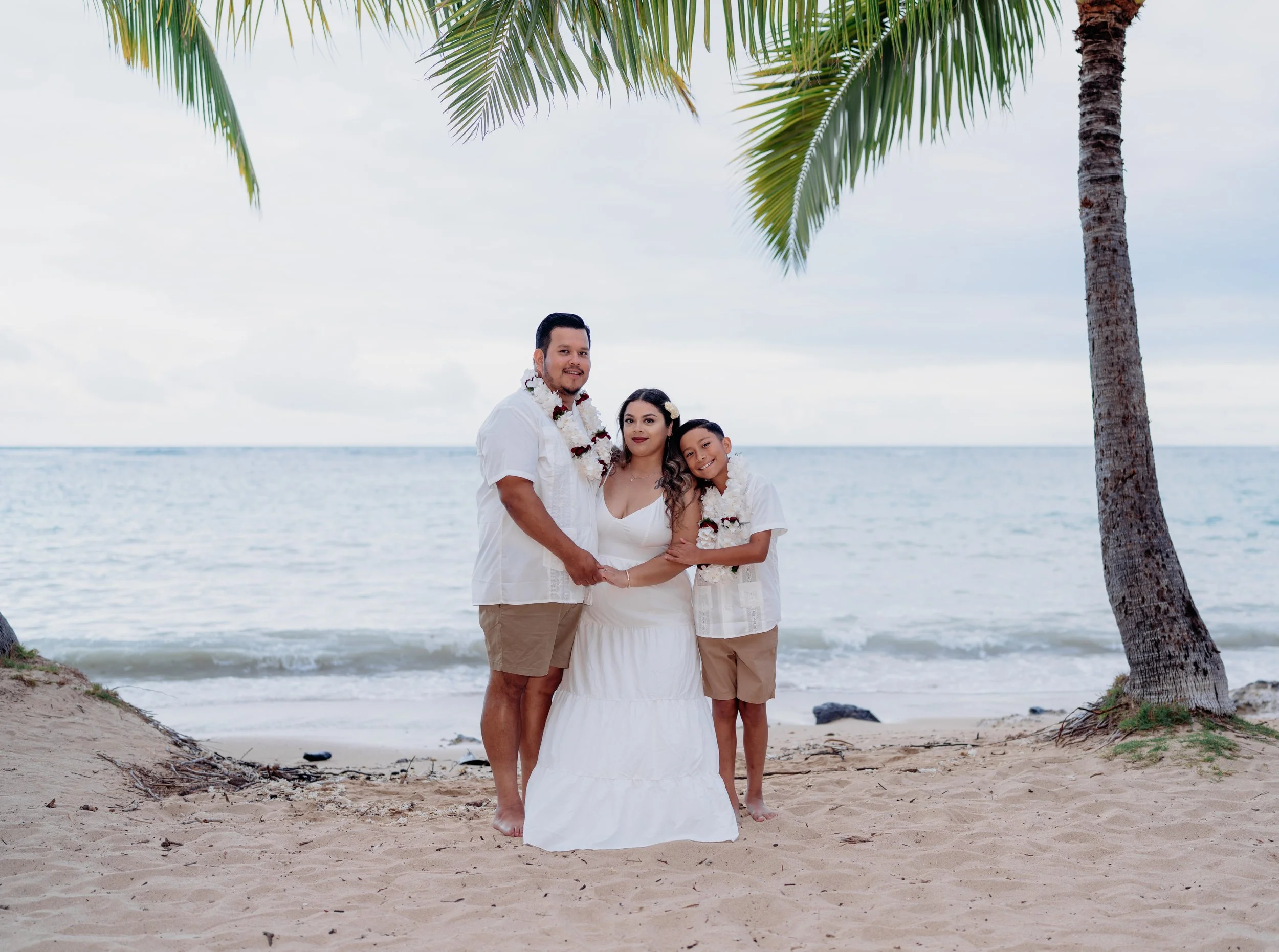 A family of three standing on a beach with sand, ocean, and palm trees in the background during daytime. The father, mother, and young son are dressed in white and tan clothing, with leis around their necks. They are smiling and holding hands.