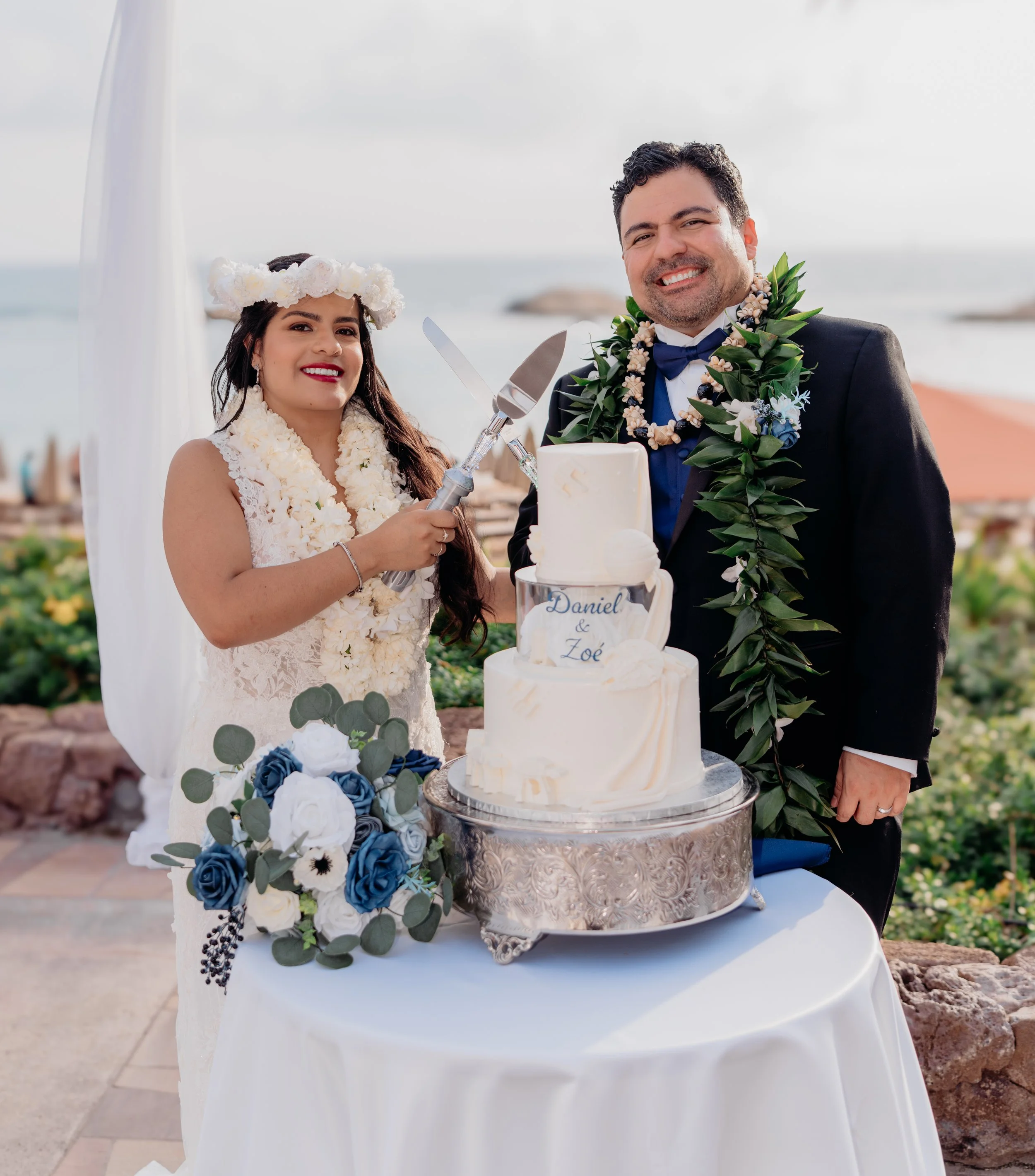 Bride and groom cutting a wedding cake outdoors by the beach, smiling, with wedding decorations and flowers in the foreground.