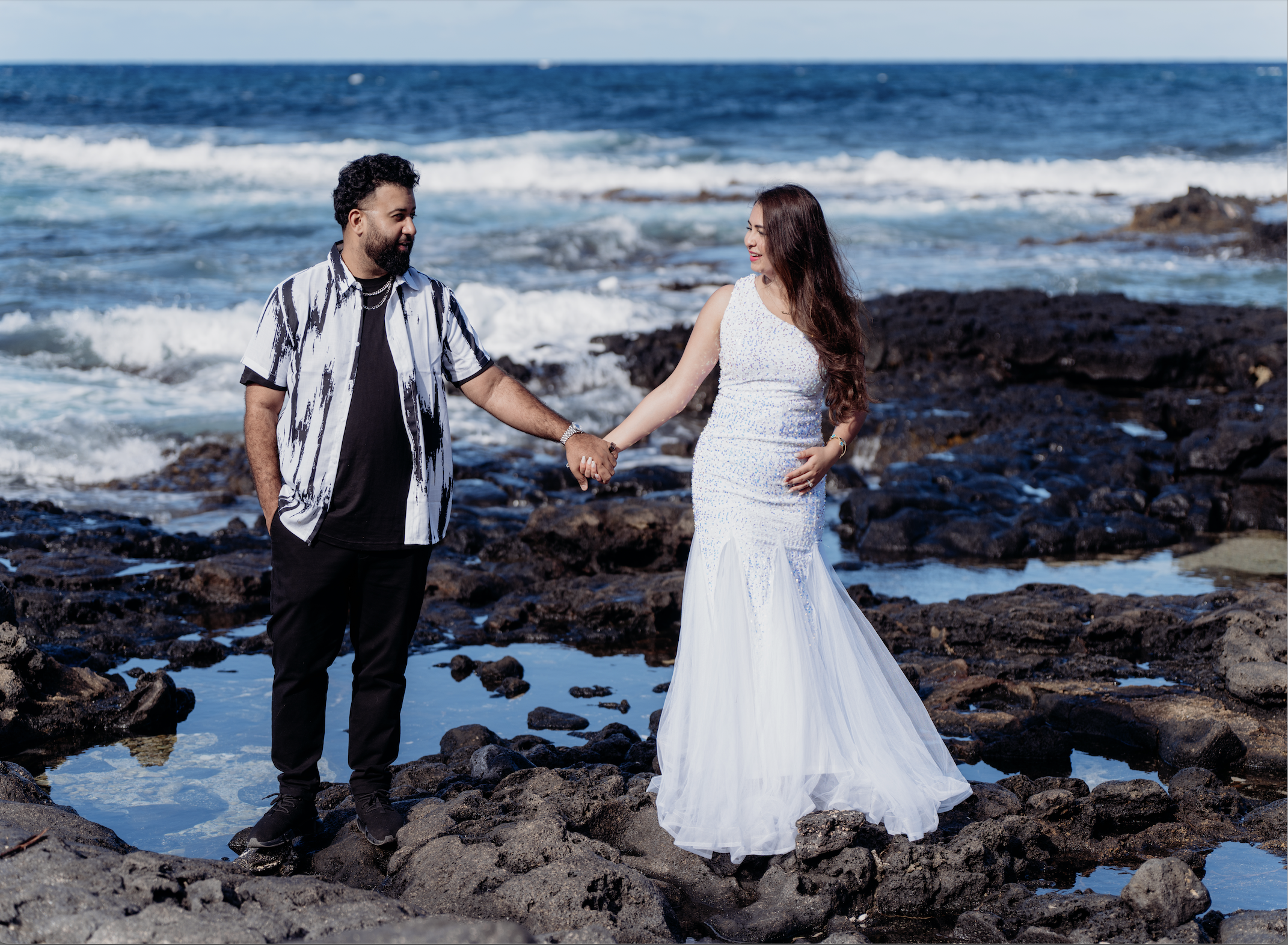 A couple holding hands on a rocky beach, with the ocean and waves in the background, dressed in wedding attire.