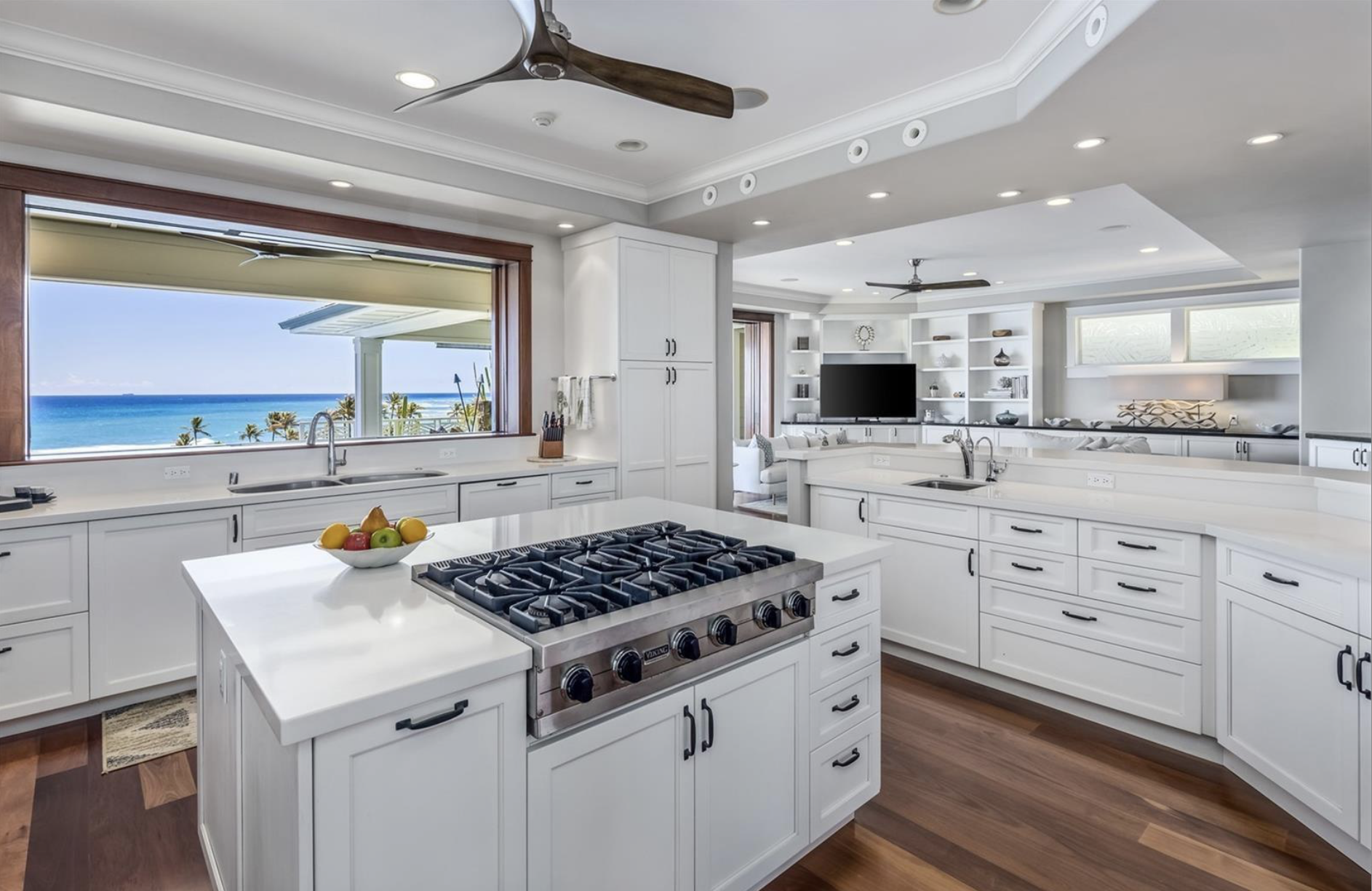 Modern white kitchen with large window showing a view of the ocean, hardwood floors, and built-in white cabinets. A kitchen island with a fruit bowl and stove is in the center. The space opens to a living area with built-in shelves and a television.