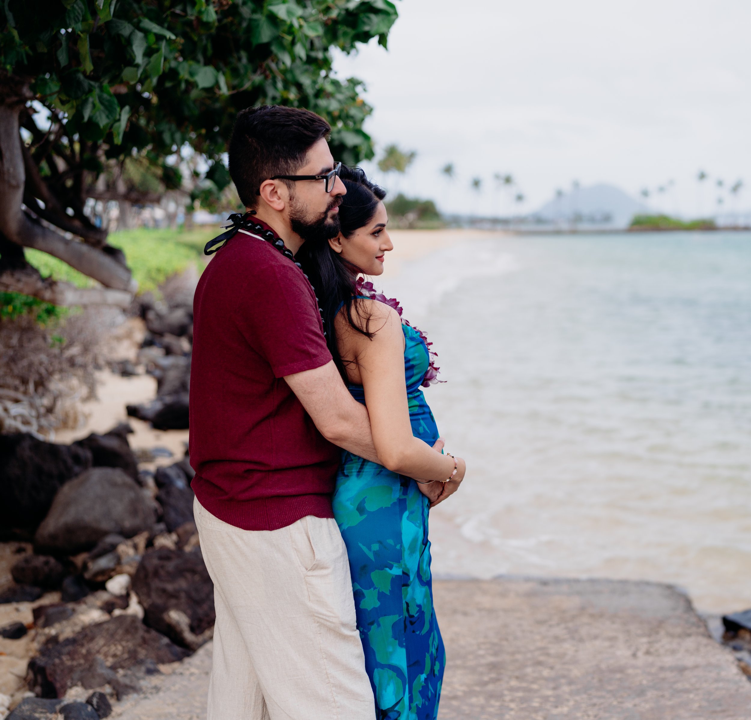 A couple stands close together on a beach, with the man behind the woman, facing the shoreline. They are under a tree with rocks along the shore, and tropical trees and water are visible in the background.