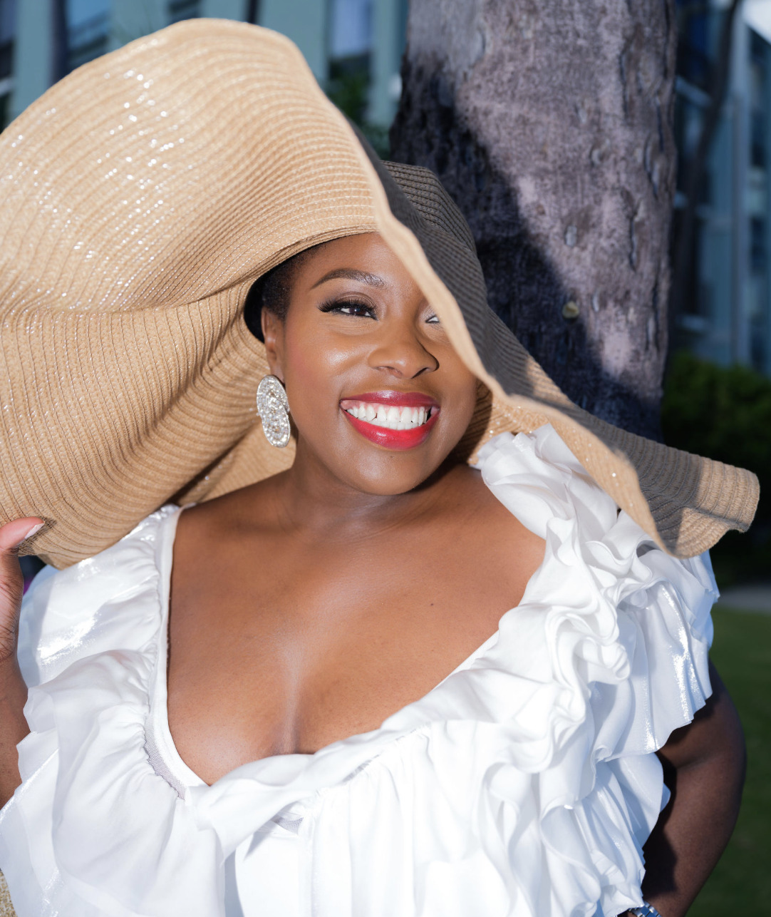 Woman smiling in a sun hat and white ruffled dress outdoors near a tree.