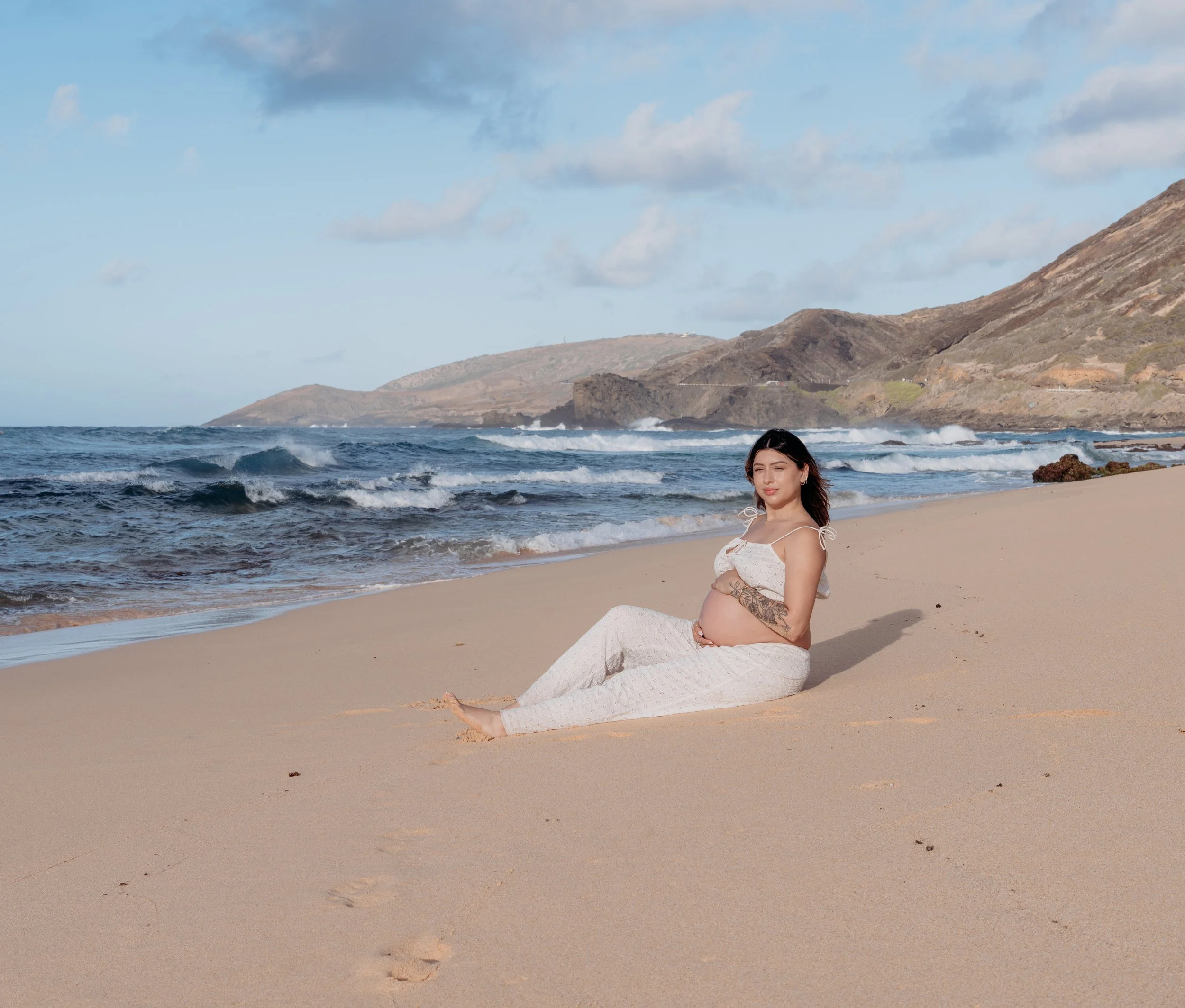 Pregnant woman sitting on sandy beach with ocean waves and hills in the background.