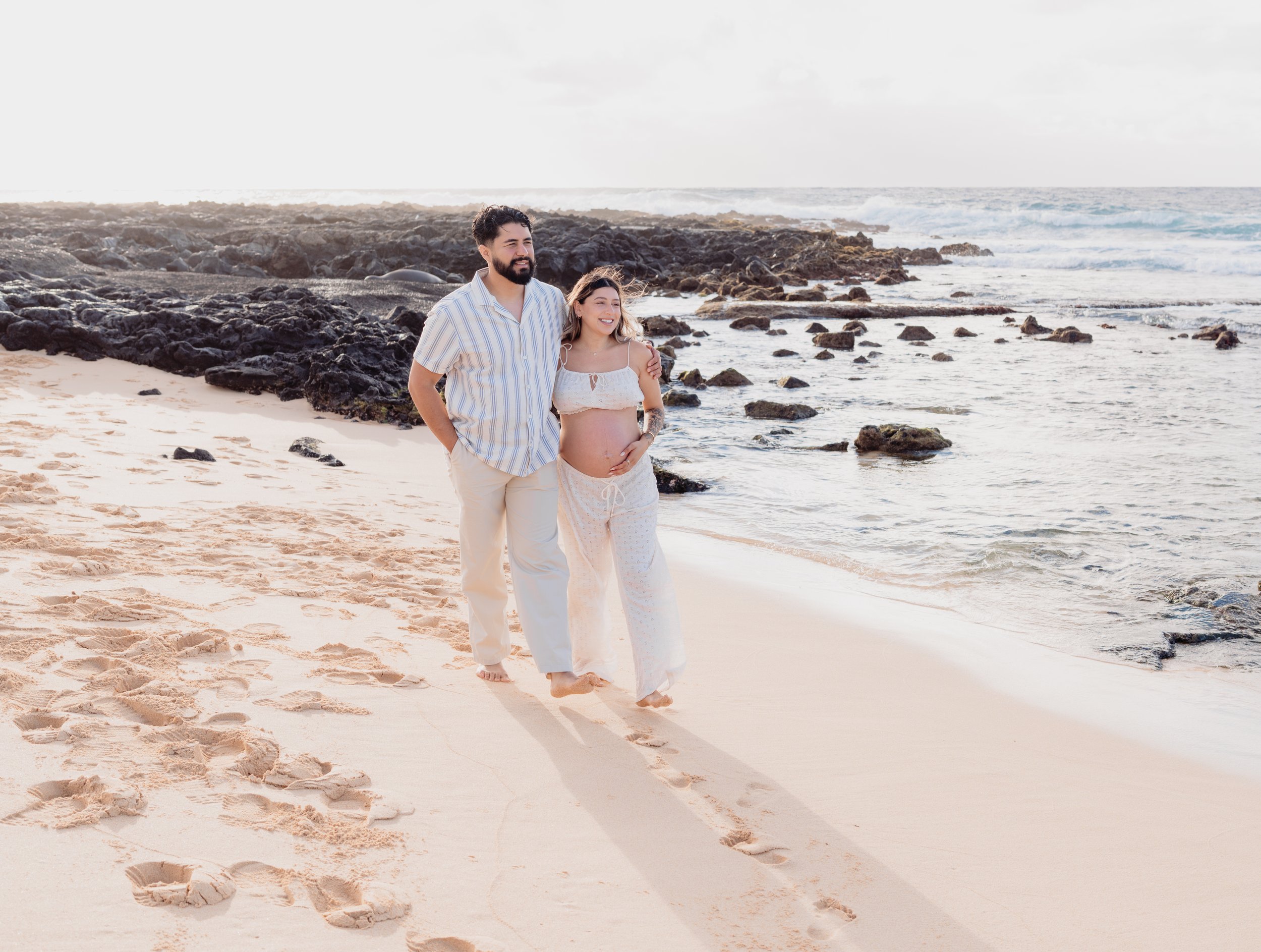 A couple walking barefoot on a sandy beach, with rocks and ocean waves in the background, during daytime.