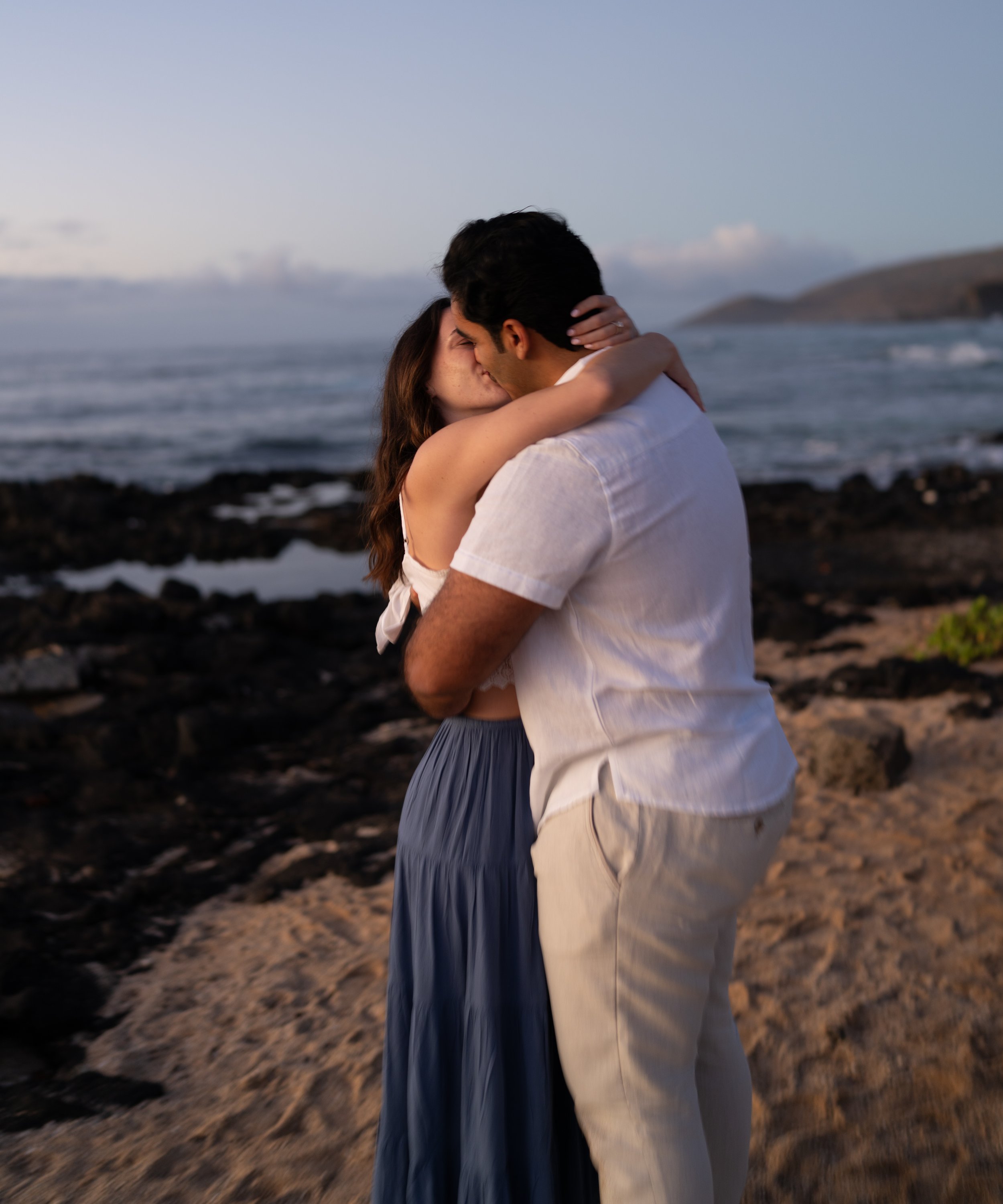 A couple kissing on the beach at sunset, with the ocean and distant hills in the background.
