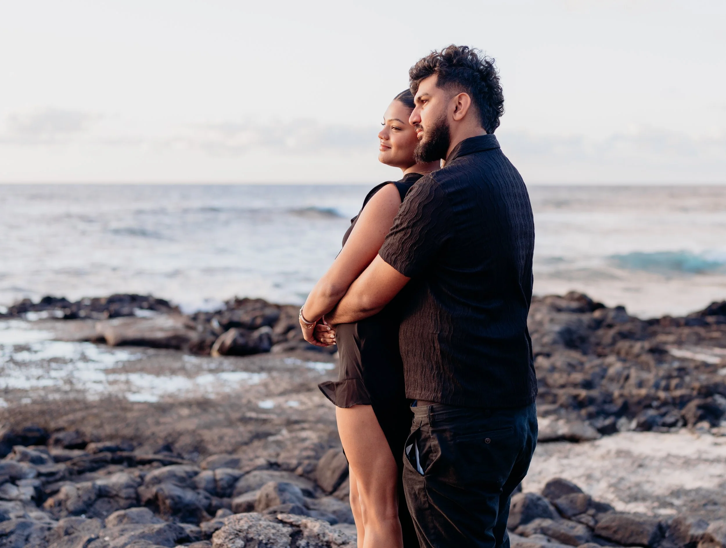 A couple stands on a rocky beach by the ocean, facing the water with their eyes closed, enjoying the peaceful moment.