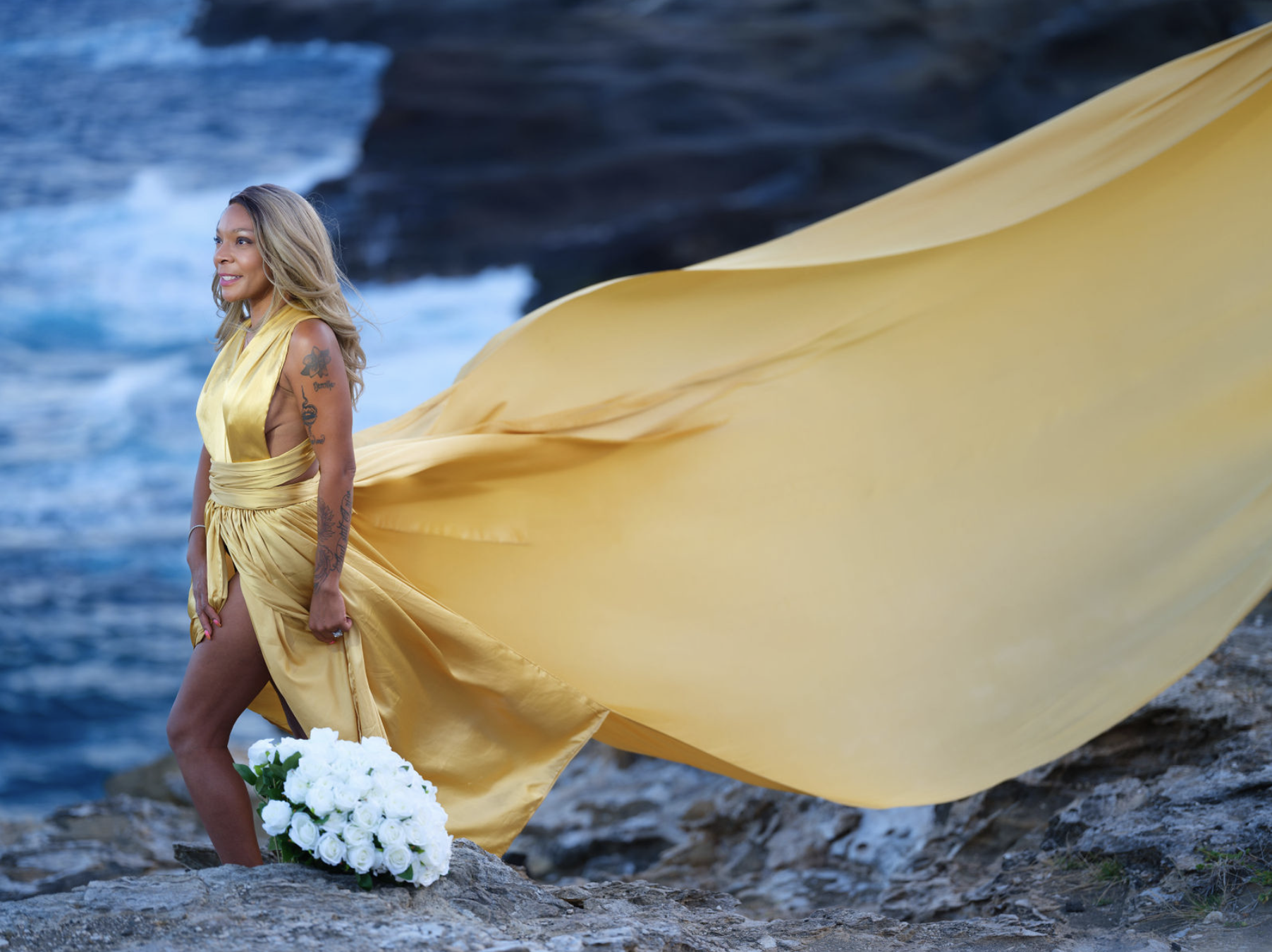 Woman in a long yellow dress holding a bouquet of white roses, standing on rocky beach with waves in the background.