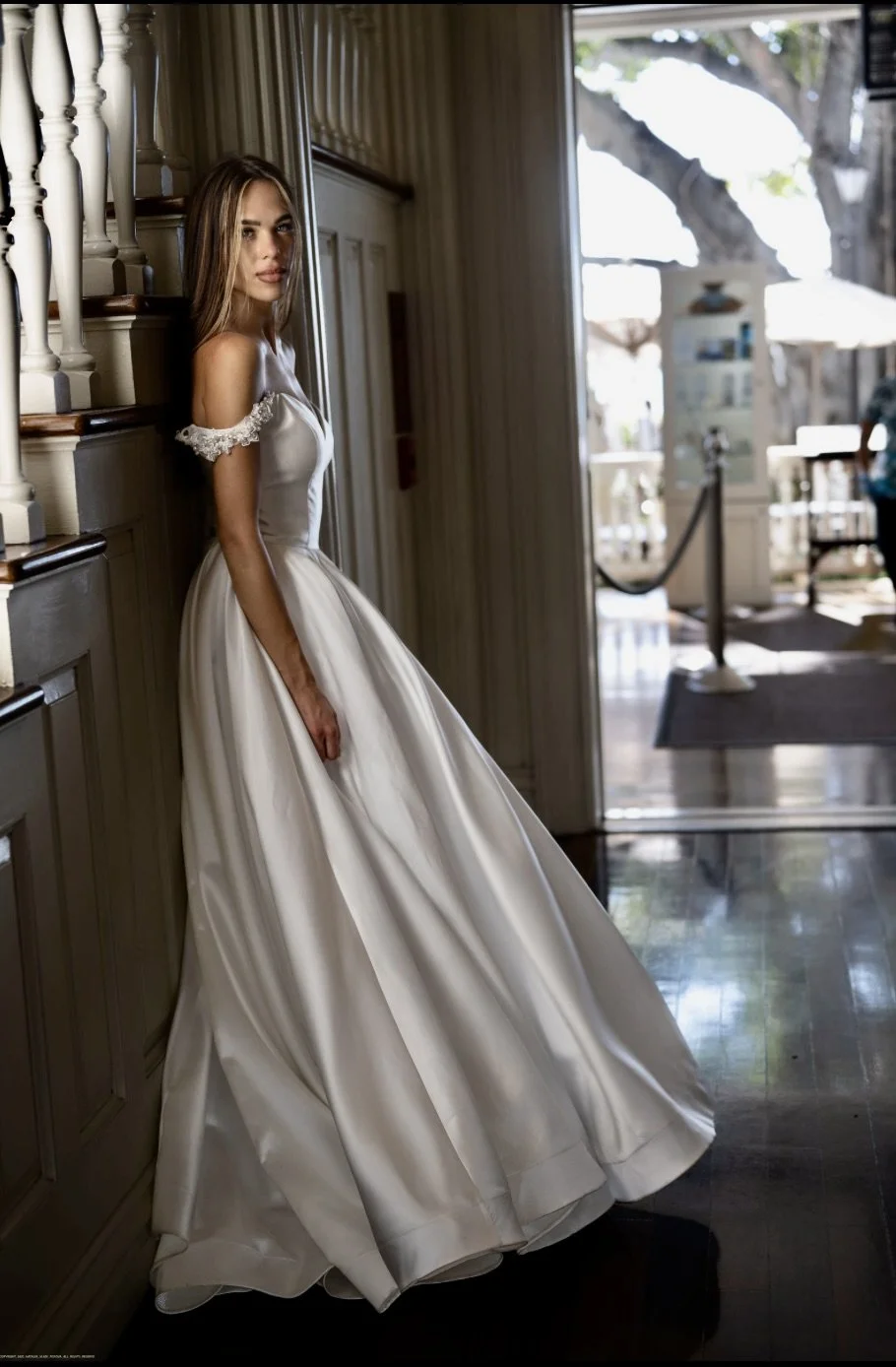 A young woman in a white wedding gown standing indoors next to a staircase, looking at the camera. There is sunlight coming through a large open door behind her, revealing an outdoor scene with trees and a patio.