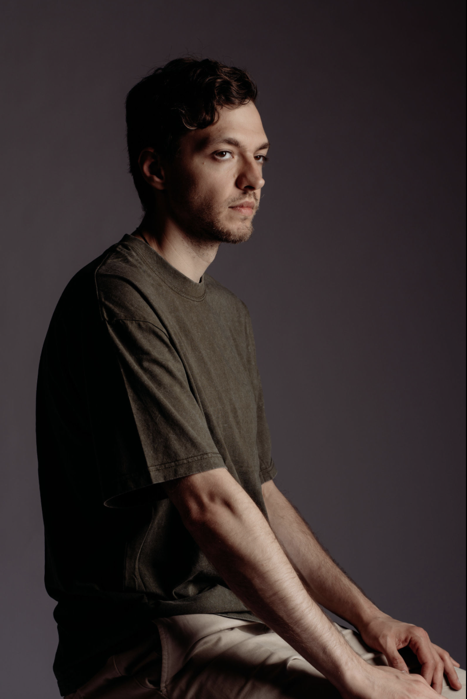 A young man with dark hair and light beard wearing a dark green T-shirt, sitting against a plain dark background, looking slightly to the side.