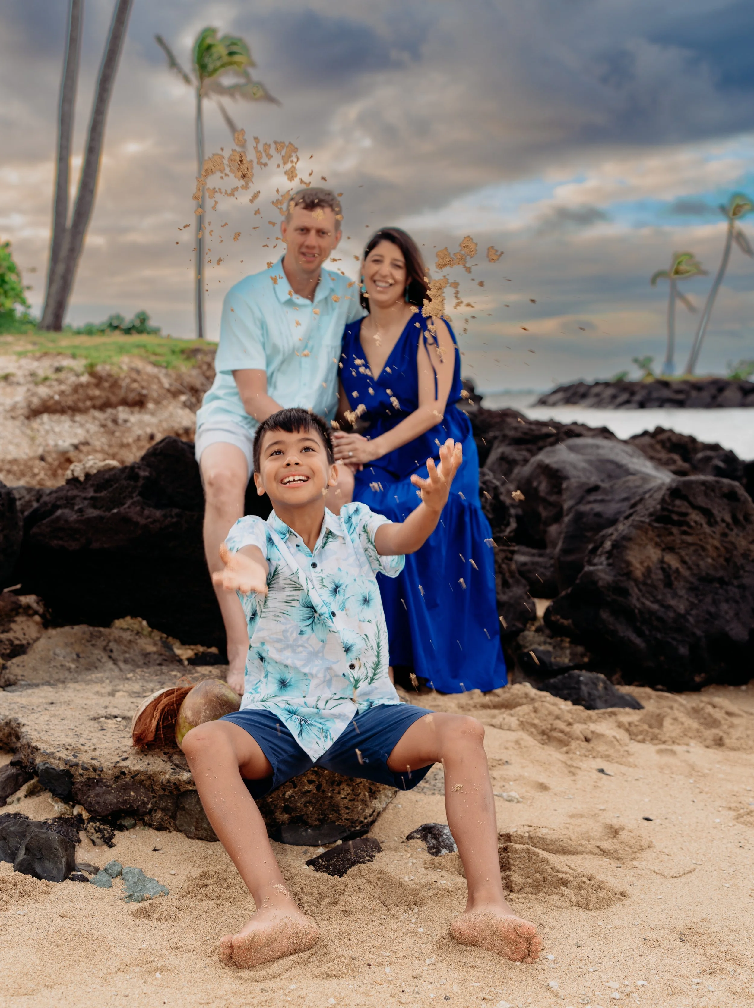 A happy family of three playing at the beach, with a young boy in the foreground splashing sand, and a man and woman sitting on rocks behind him smiling. The sky is partly cloudy with palm trees in the background.
