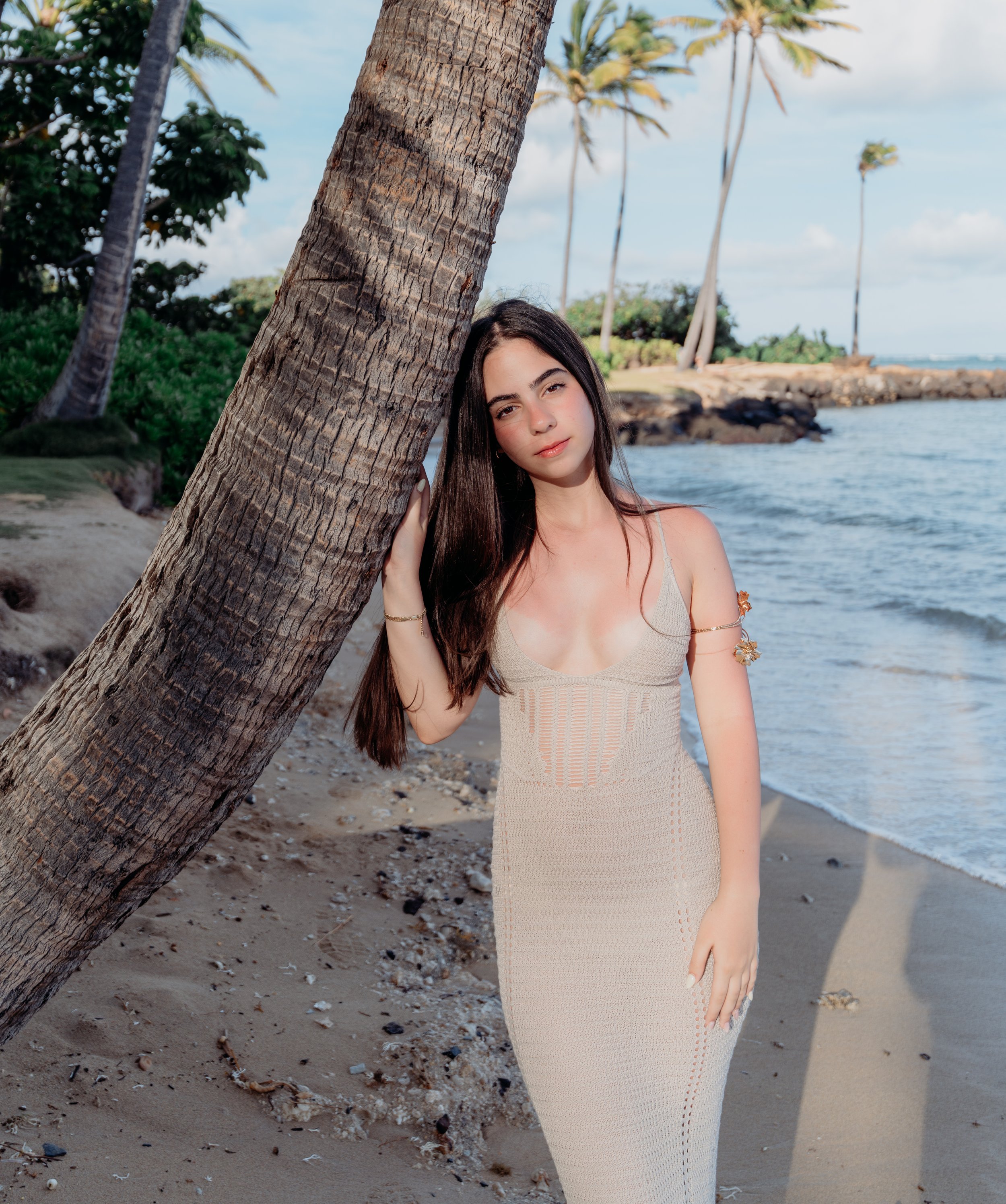 A young woman with long dark hair in a beige dress standing next to a leaning palm tree on a sandy beach near the ocean, with trees and a blue sky in the background.