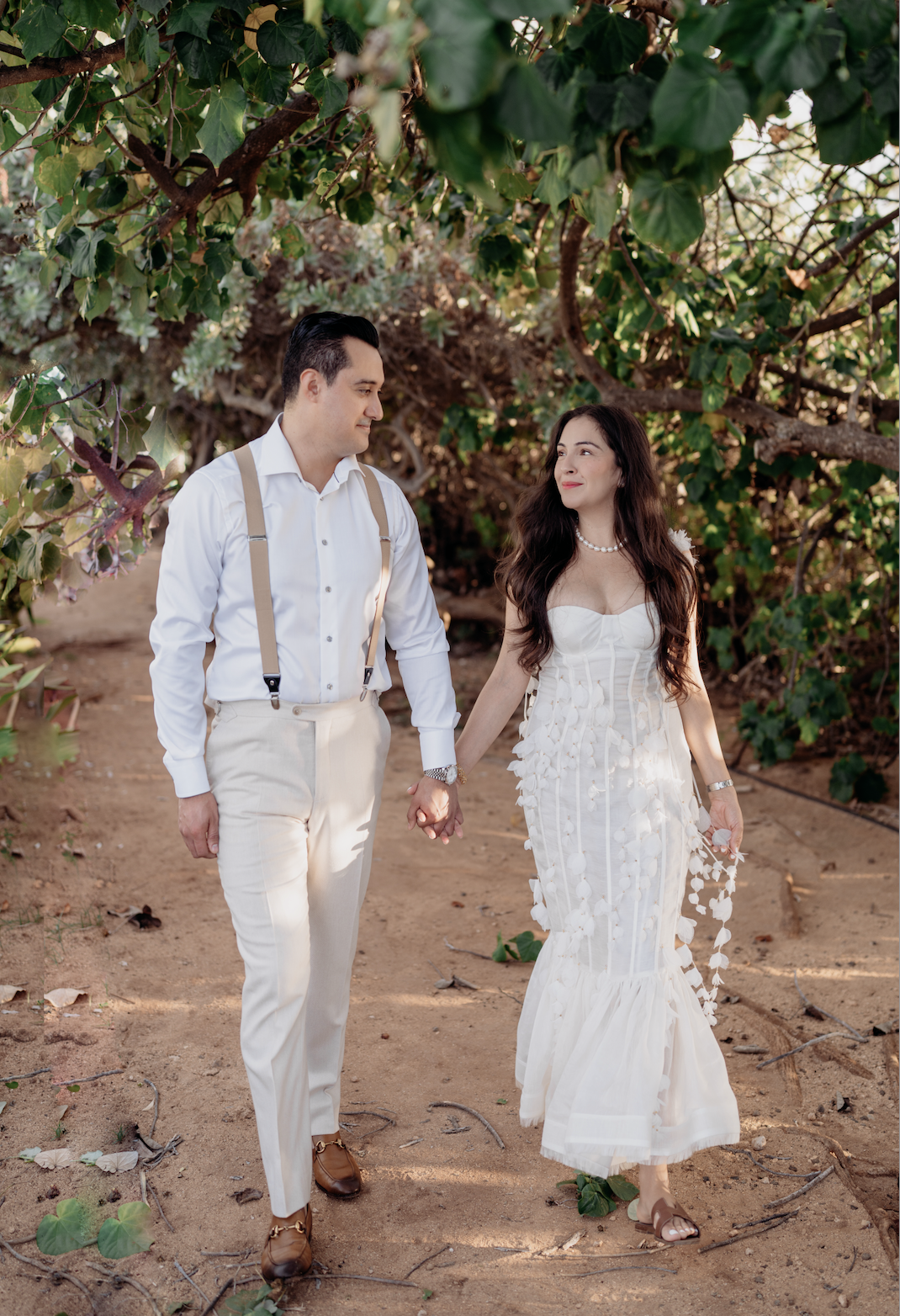 A couple holding hands walking on a dirt path surrounded by green foliage, dressed in elegant white clothes, with the woman wearing a white dress and the man wearing a white shirt with suspenders.