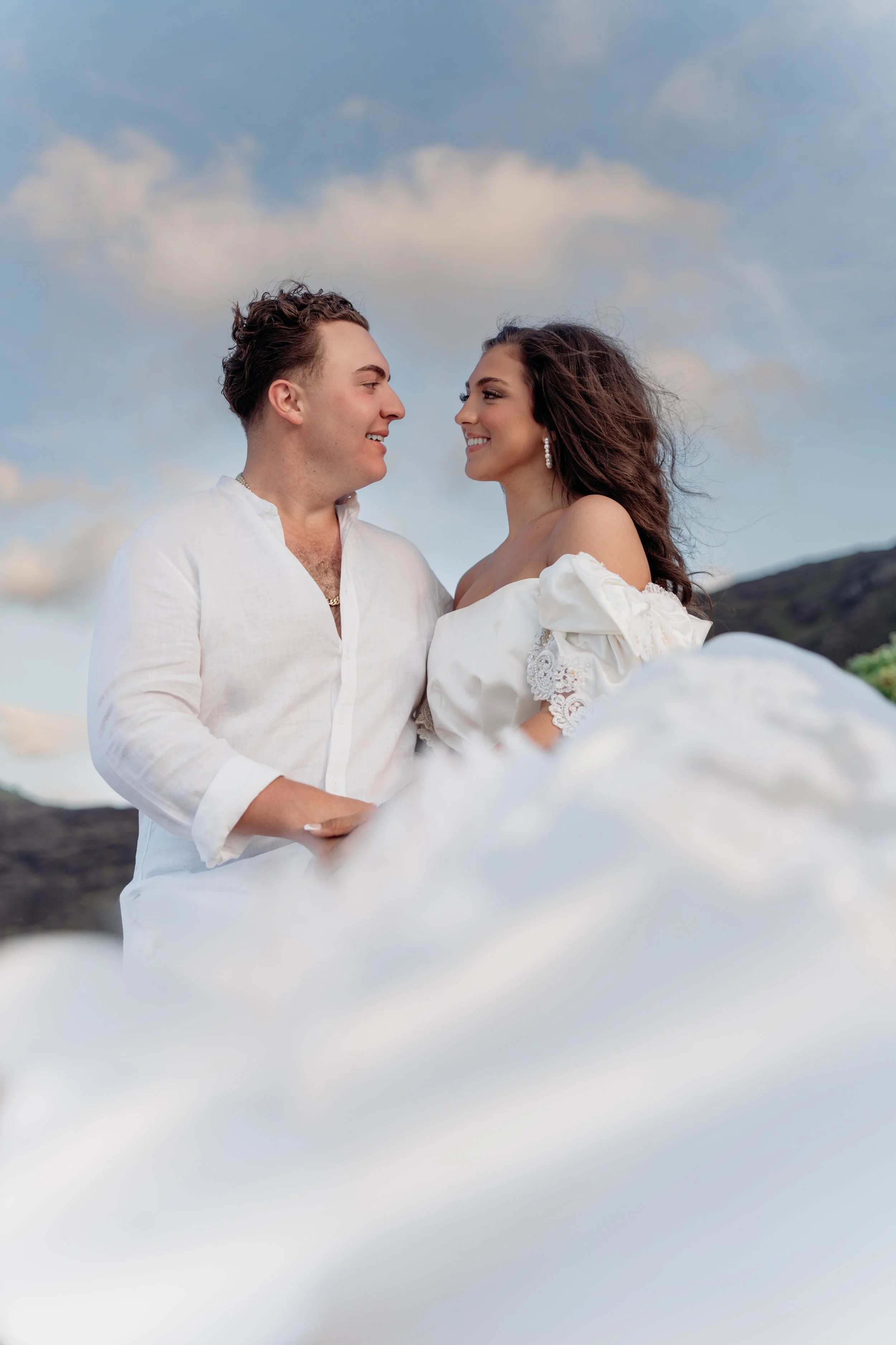 A couple smiling and gazing at each other outdoors with clouds and sky in the background, dressed in white, standing behind clouds or mist.